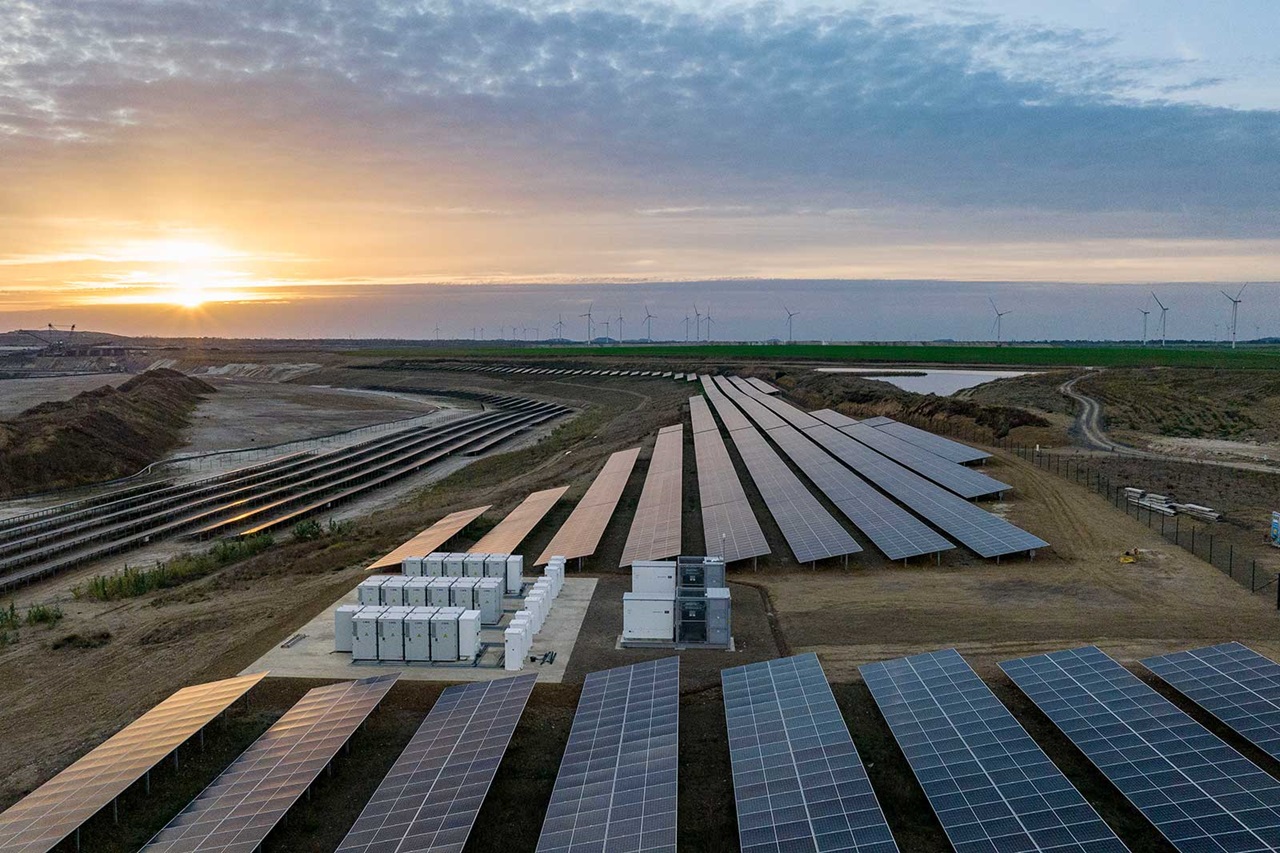 Solarpaneele in einer weitläufigen Landschaft bei Sonnenuntergang, mit Windkraftanlagen im Hintergrund.