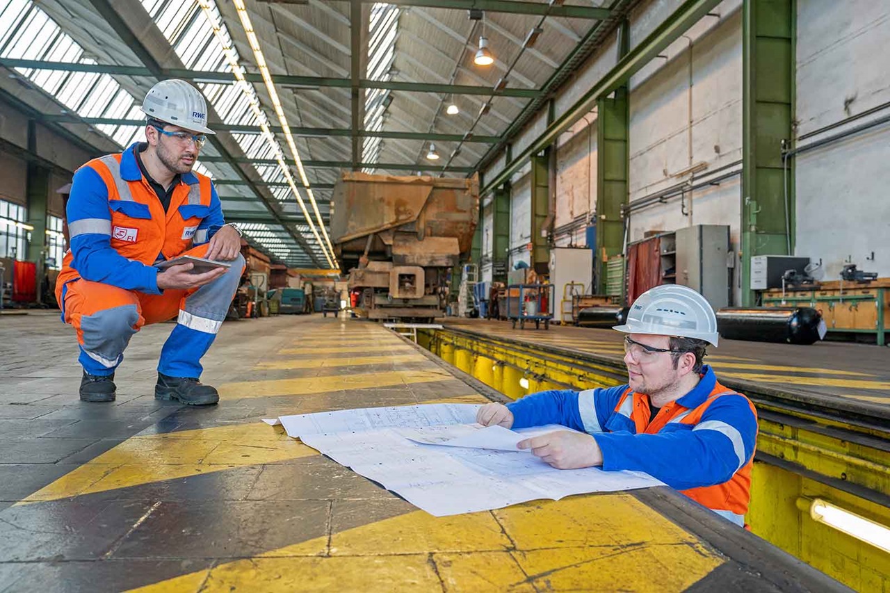 Zwei Arbeiter in Schutzkleidung besprechen Pläne auf einem großen Blatt Papier in einer geräumigen Industriehalle.