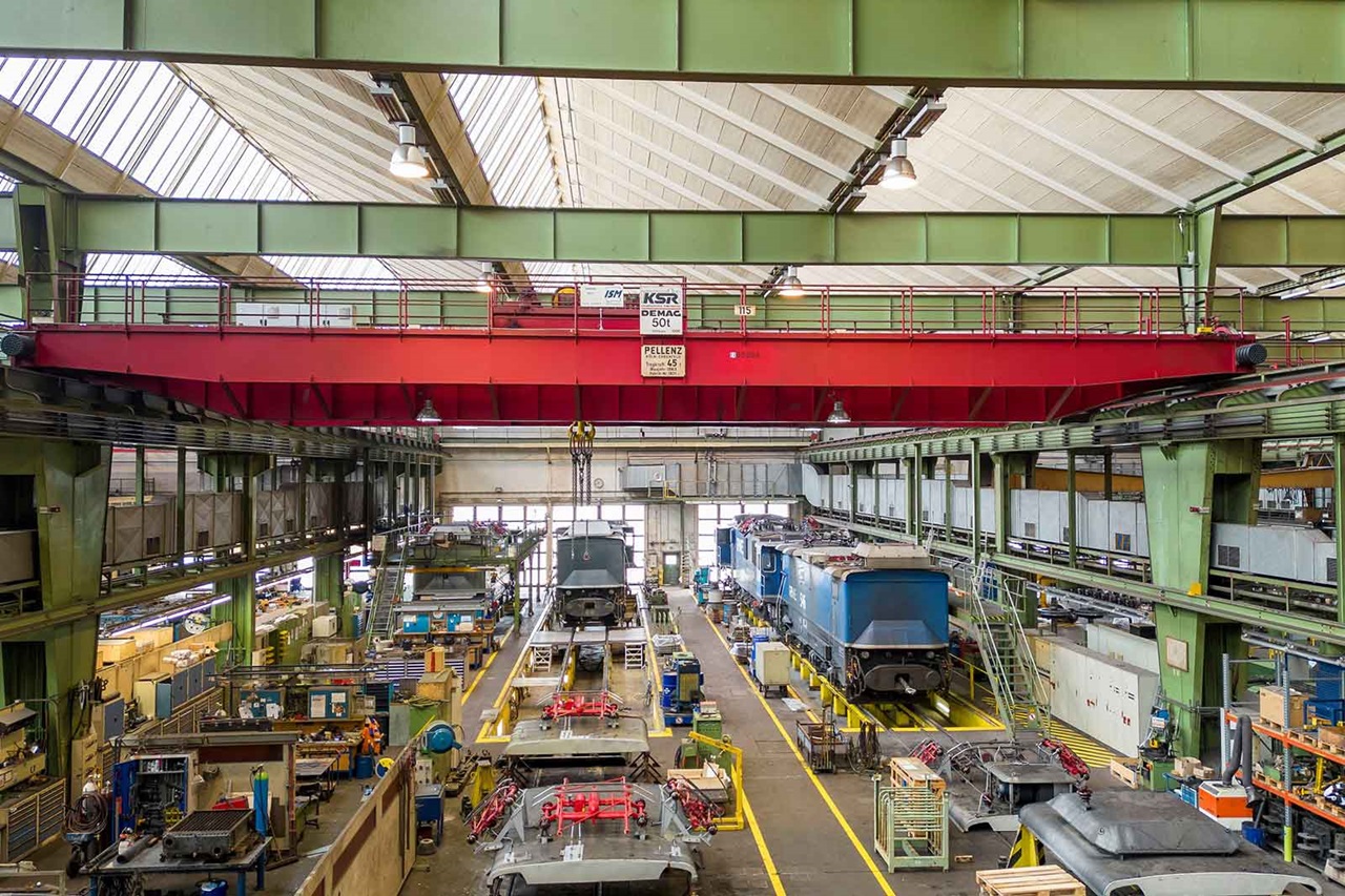 Eine Industriewerkstatt mit Oberkranen und verschiedenen Zügen, die unter einem großen Dach mit Oberlichtern gewartet werden.