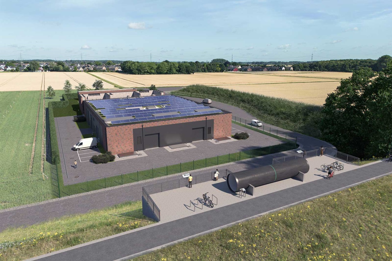 Ein rotes Backsteingebäude mit Solarpaneelen auf dem Dach steht neben Ackerland; ein eingezäunter Vorplatz, eine große schwarze Röhre, Fahrradständer und Personen sind zu sehen.