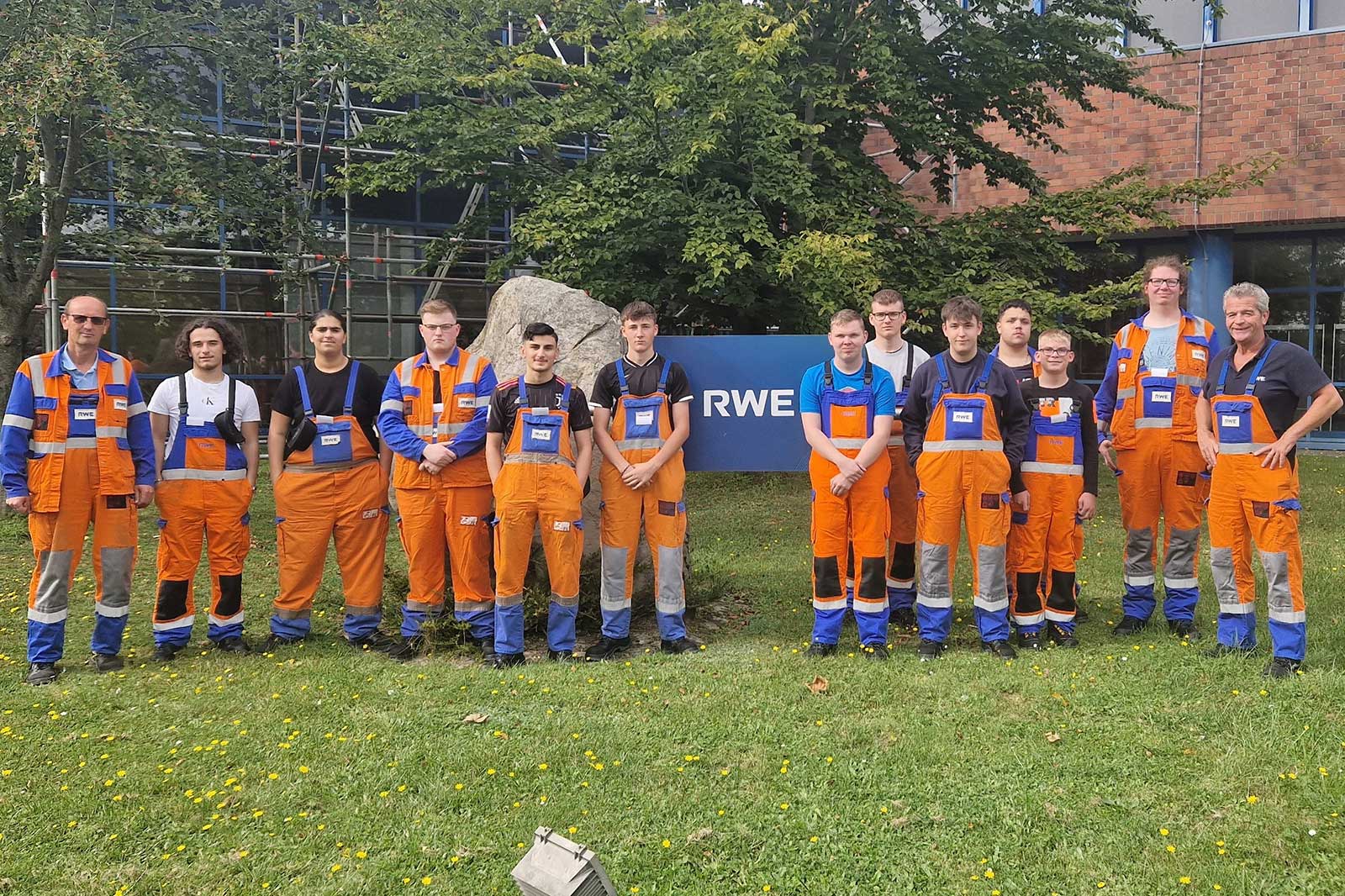 Eine Gruppe von Arbeitern in leuchtend orangefarbenen Overalls steht in einem Grasbereich neben einem Gebäude mit einem RWE-Schild.
