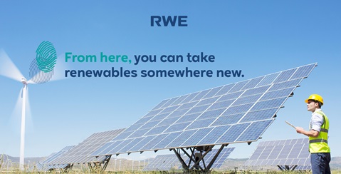 A person in a hard hat inspects solar panels with a wind turbine in the background against a clear blue sky.
