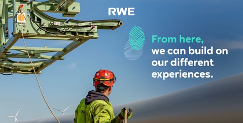 A worker in a red helmet and green jacket stands before a modern wind turbine on a construction framework.