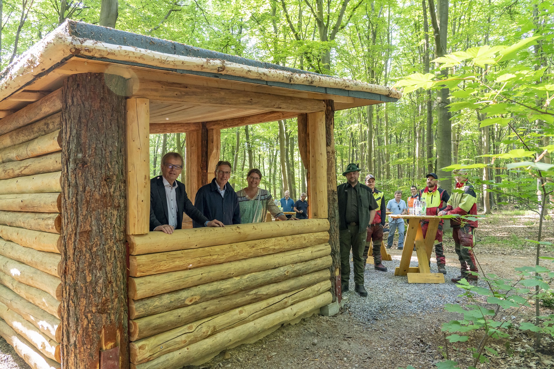 Holzhütte im Wald, Menschen versammeln sich um einen Tisch, umgeben von Grün.