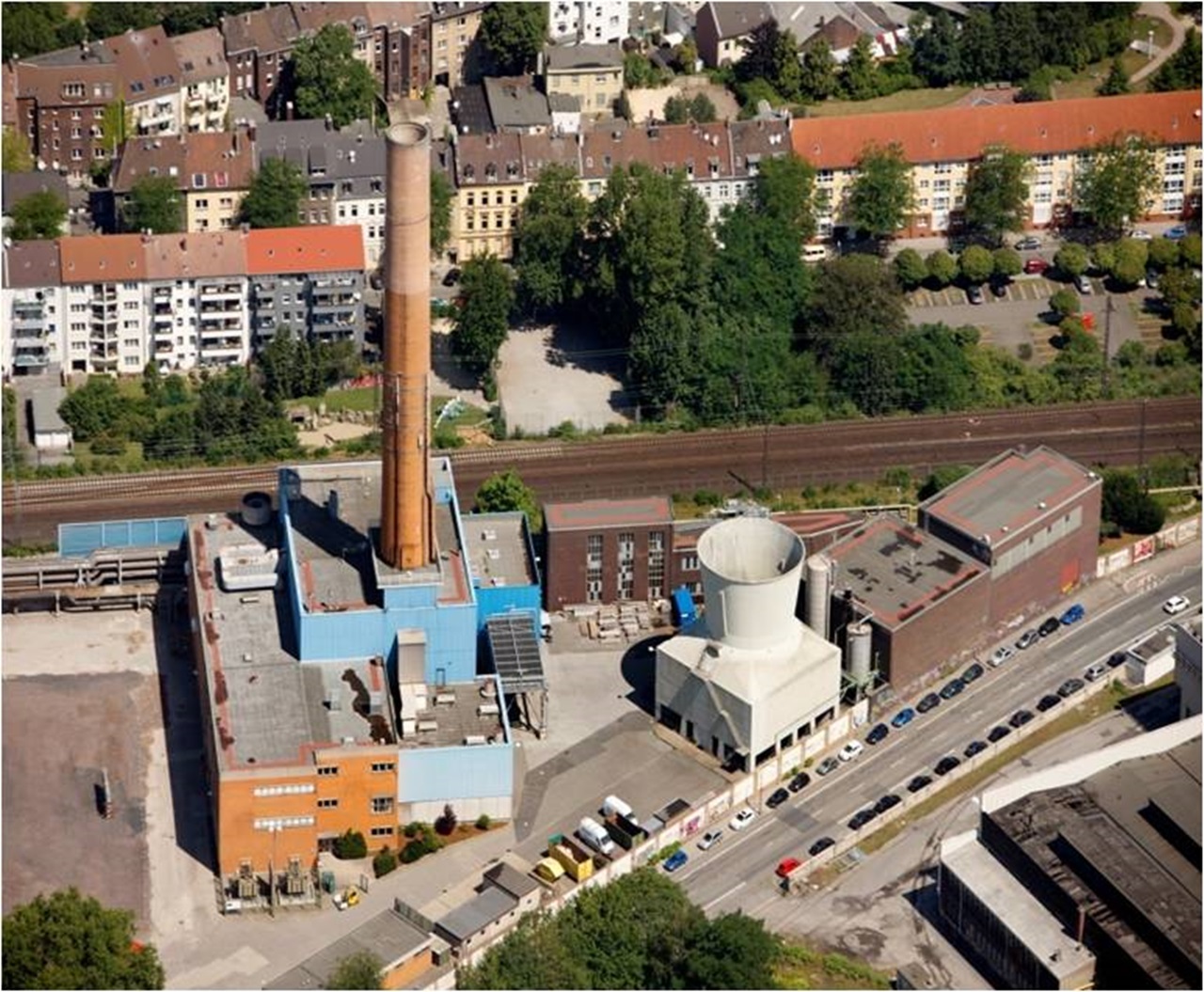 Luftaufnahme eines Industriegeländes mit einem hohen Schornstein, einem Kühlturm und umliegenden Wohngebäuden.