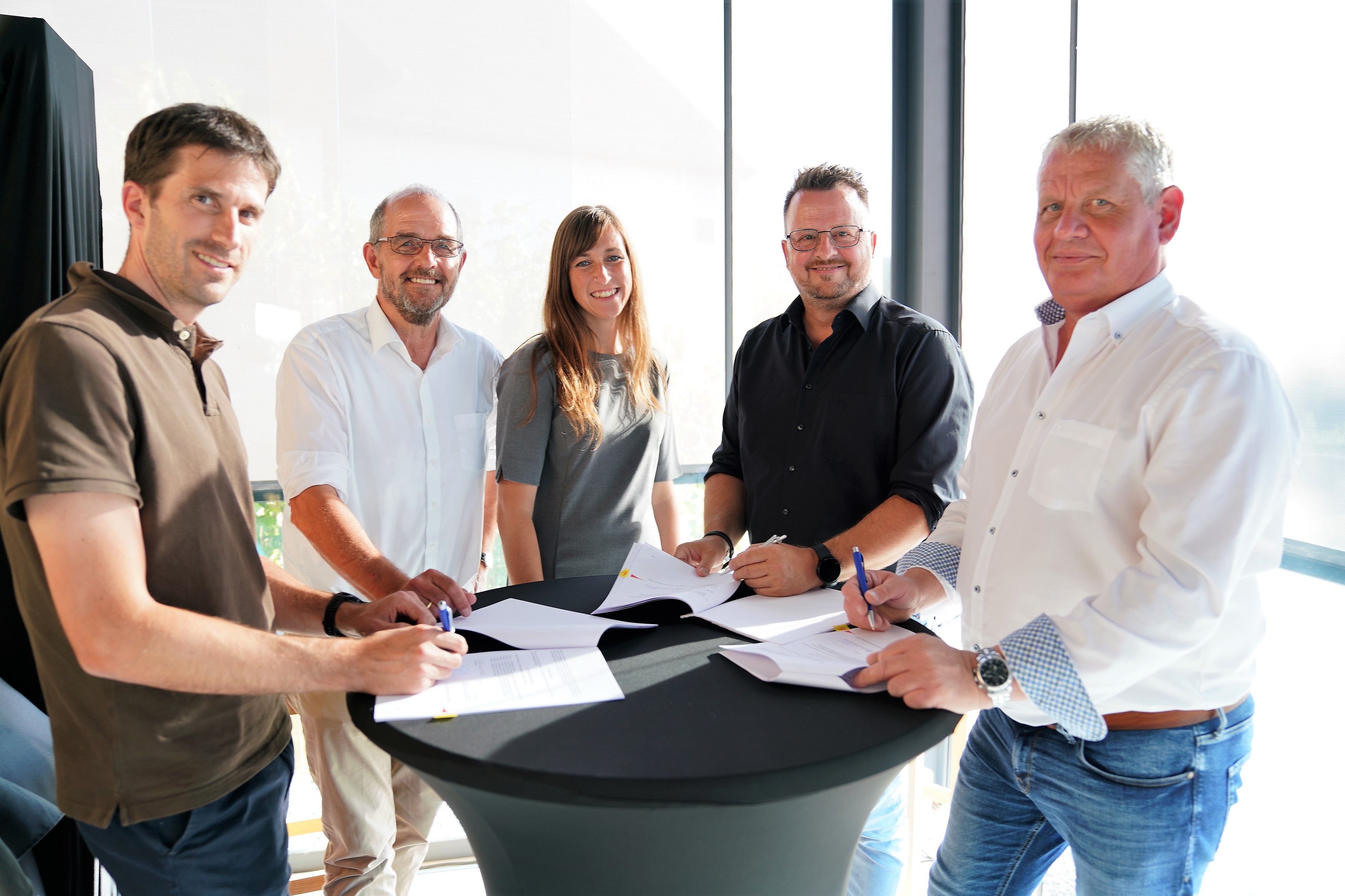 Eine Gruppe von Personen unterzeichnet Dokumente an einem runden Tisch in einem sonnigen Innenraum.