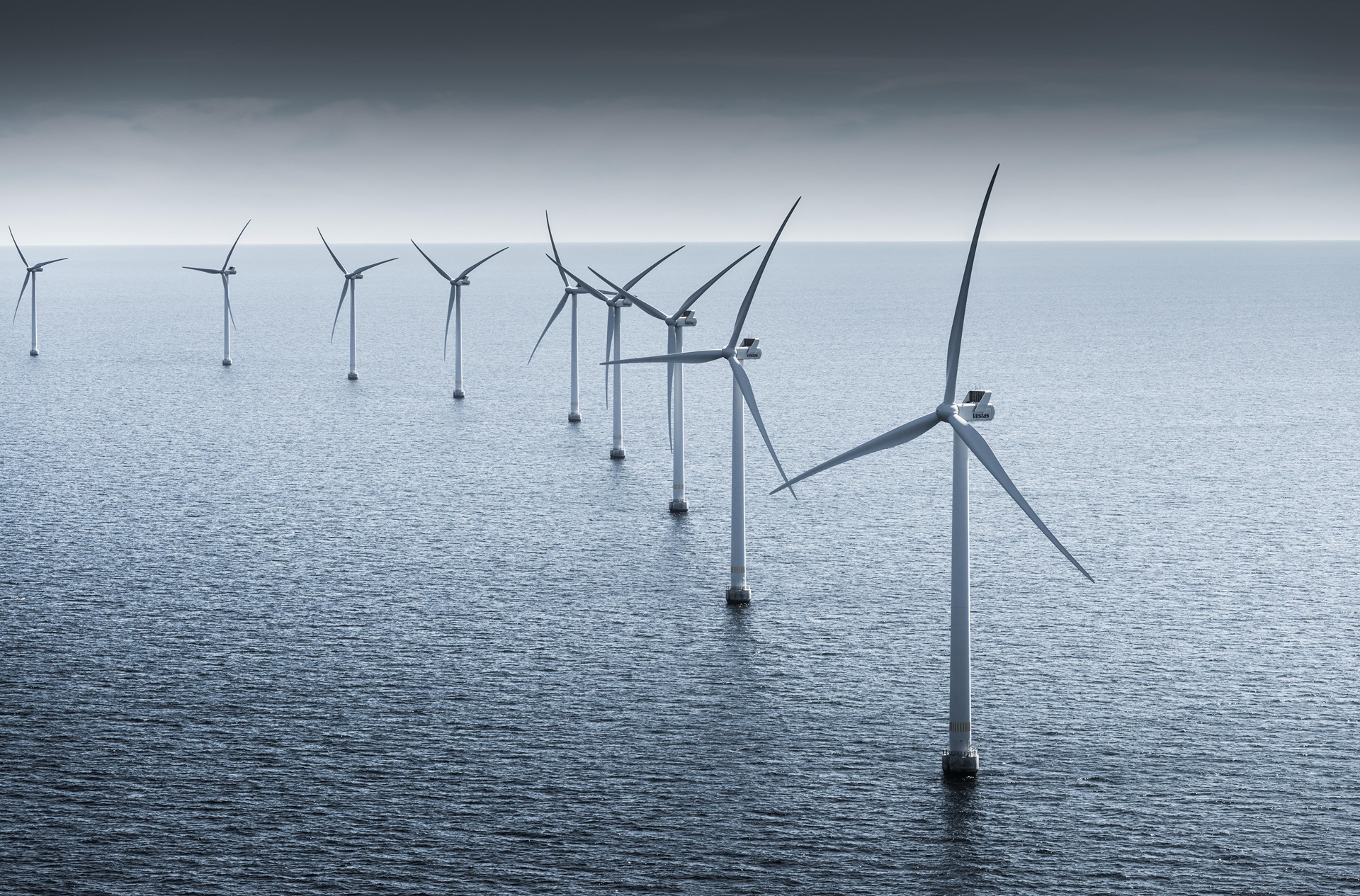 A row of offshore wind turbines standing in calm waters, under a grey sky.