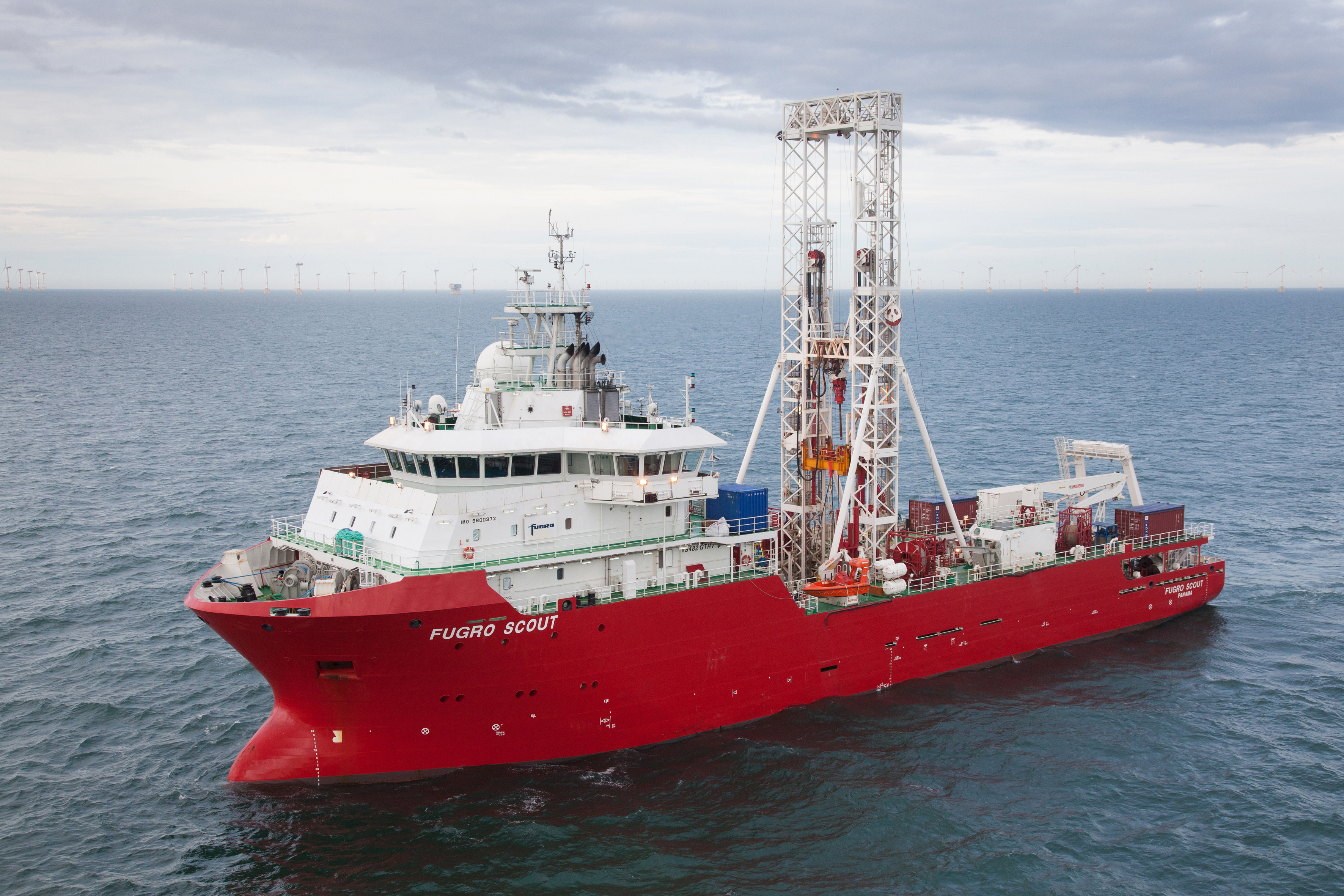 A red research vessel named Fugro Scout on the ocean, with wind turbines visible in the background.