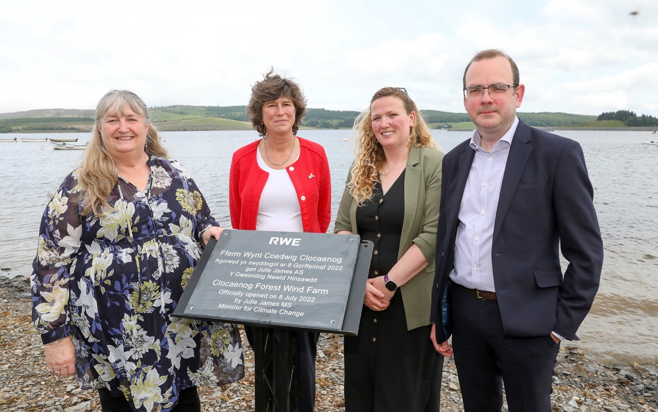 A group stands by a lake, unveiling a sign for the Clocaenog Forest Wind Farm. It is cloudy and green hills are in the background.