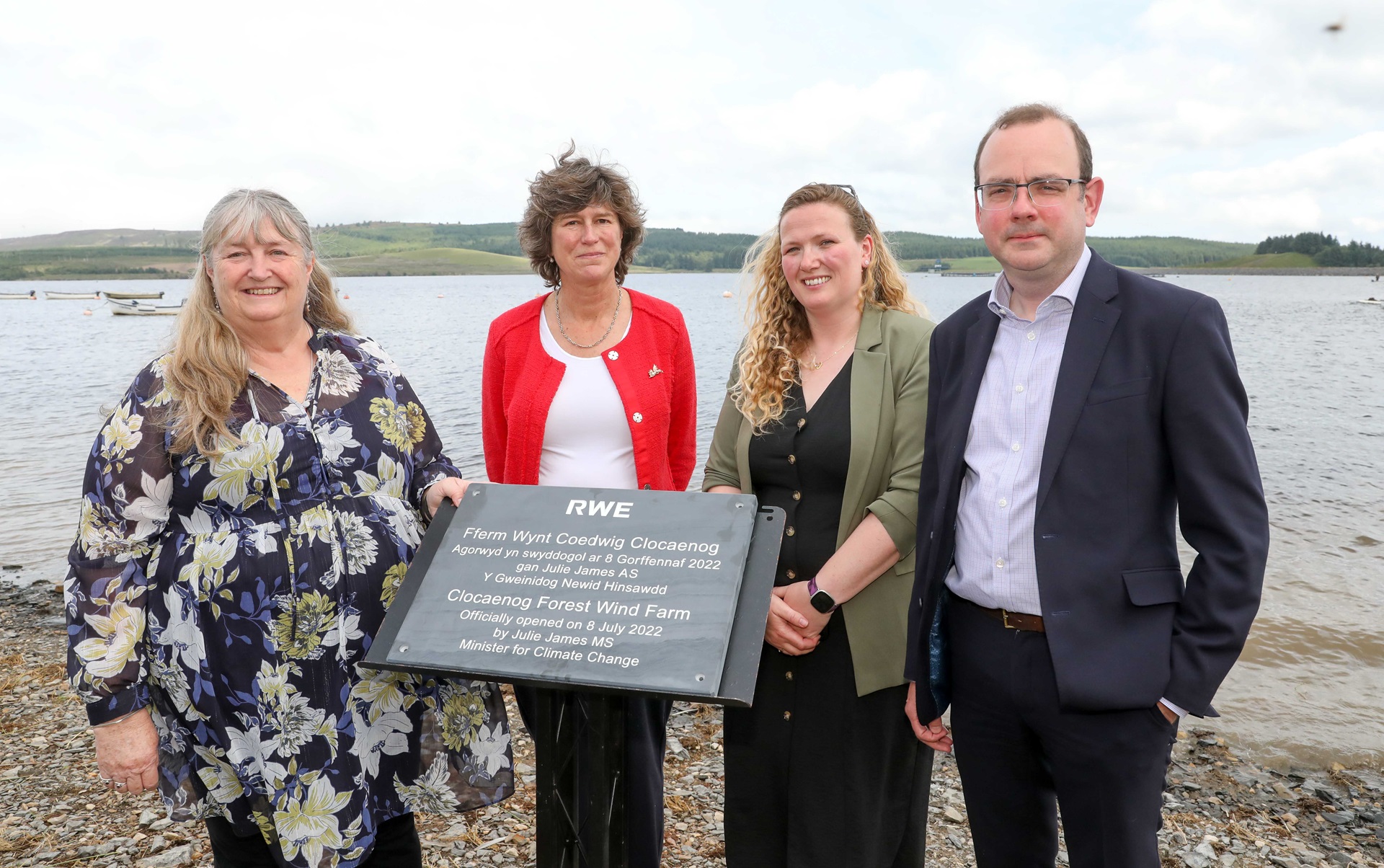 A group stands by a lake, unveiling a sign for the Clocaenog Forest Wind Farm. It is cloudy and green hills are in the background.