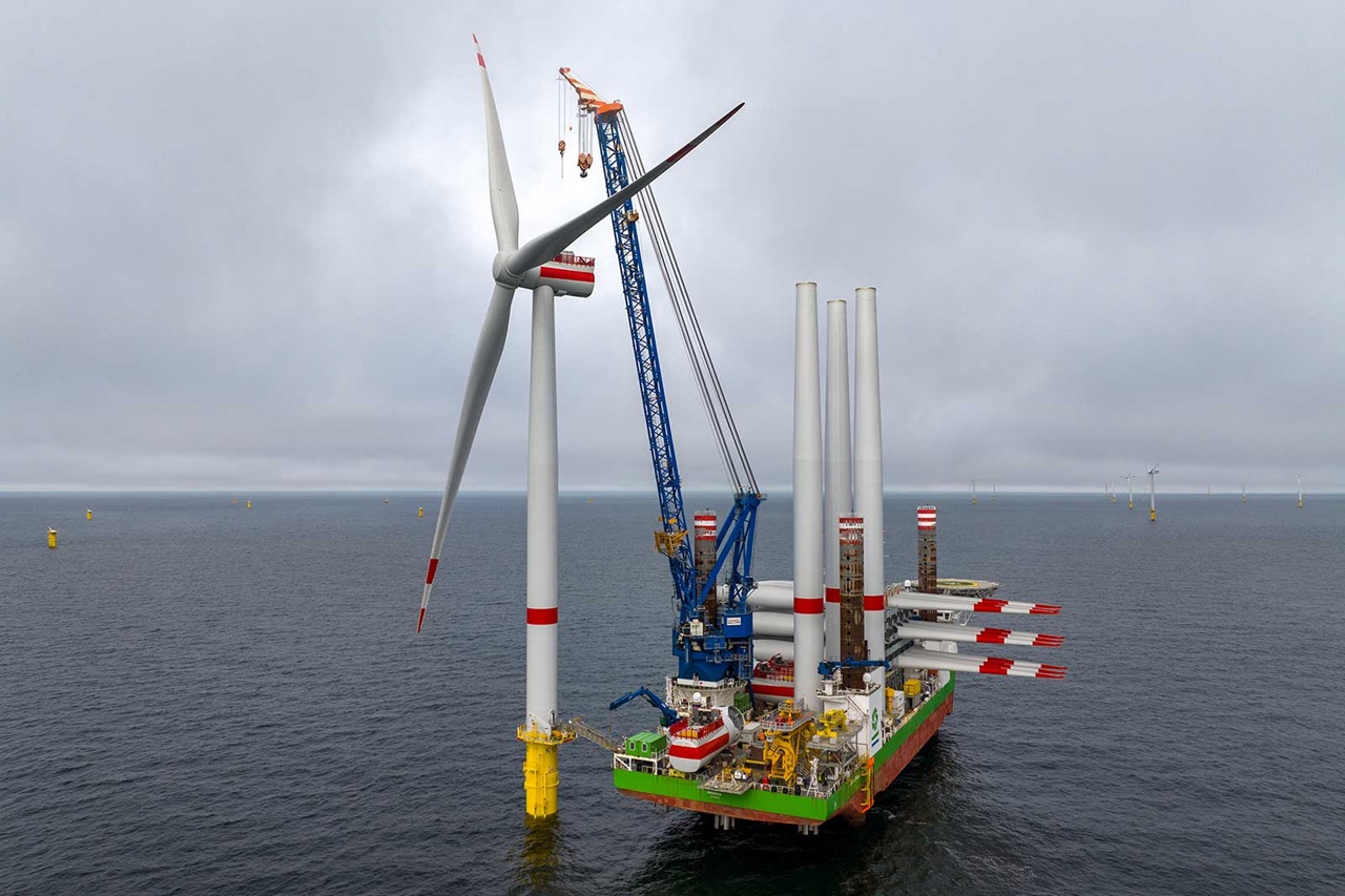 The image shows a ship with a crane installing a wind turbine at sea under a grey sky.