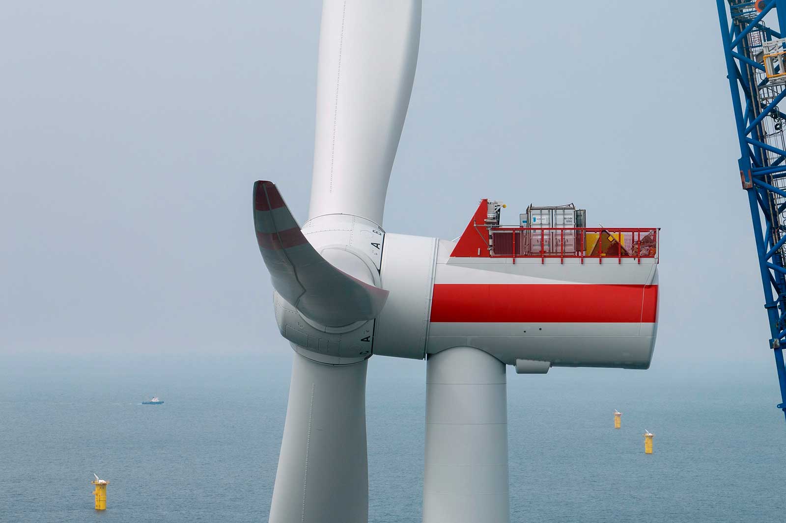 An offshore wind turbine with a red and white design, surrounded by ocean and yellow buoys in the background.