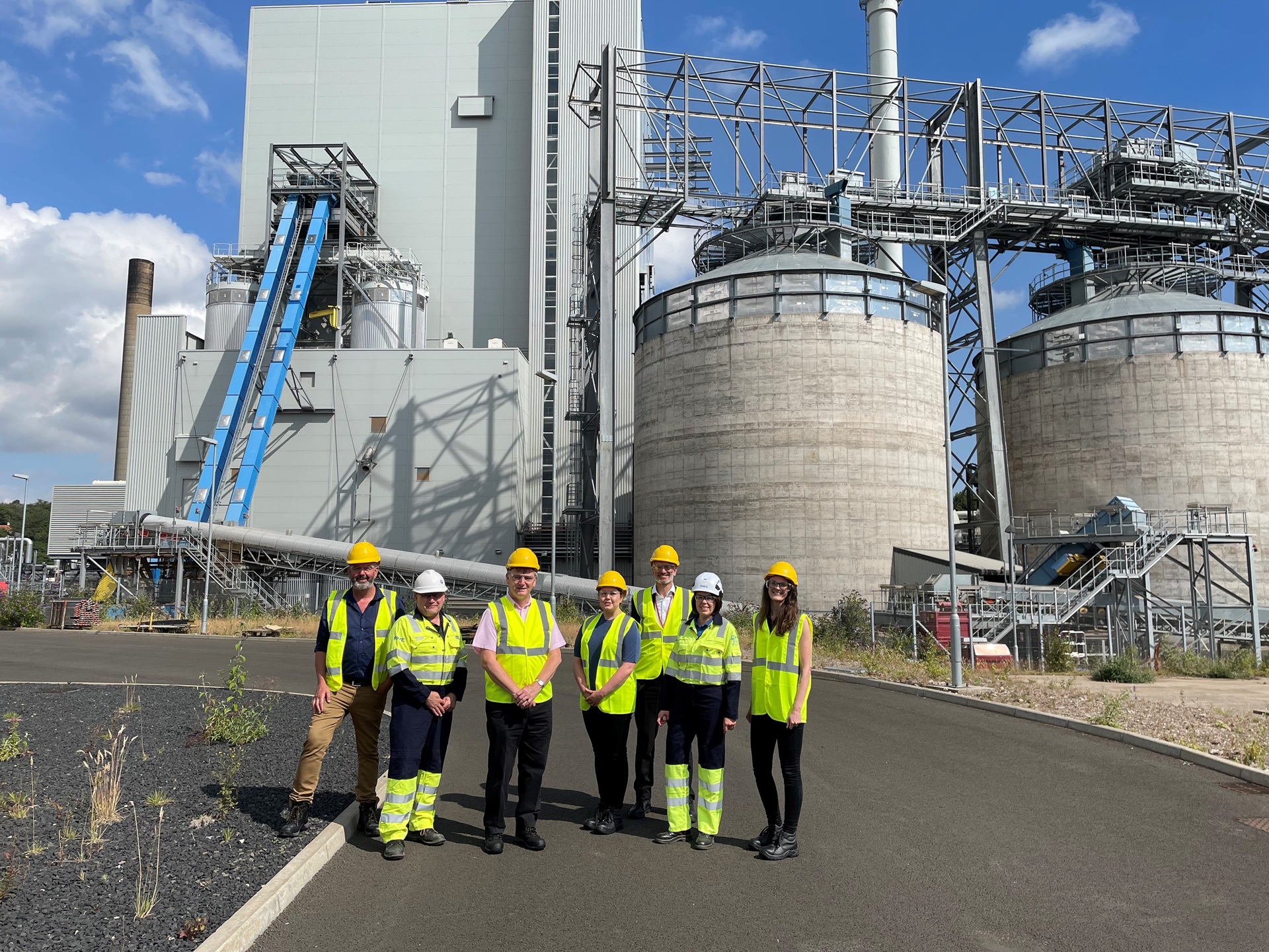 A group of people in yellow vests and hard hats in front of an industrial building with large tanks.