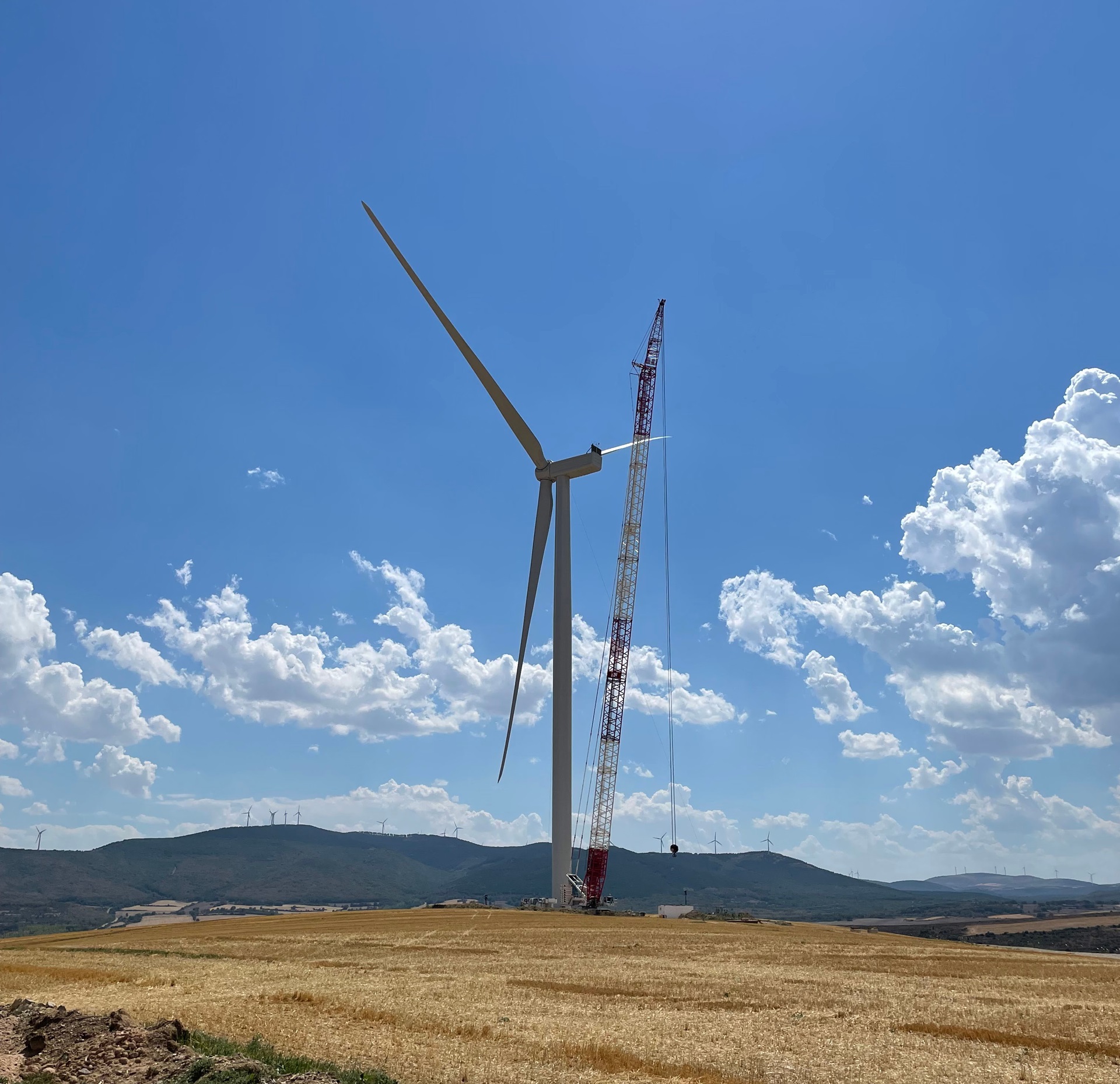 Eine im Bau befindliche Windturbine mit einem großen Kran auf einem Feld. Blauer Himmel mit Wolken im Hintergrund.