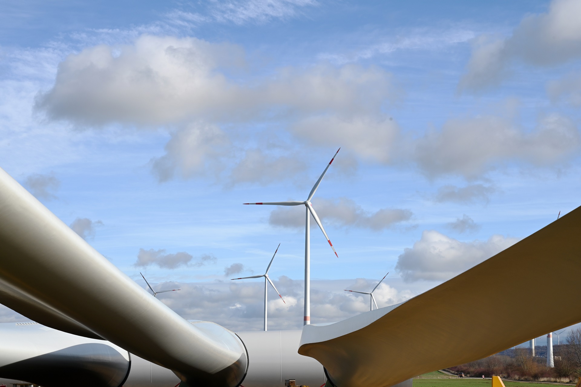 Windturbinen im Hintergrund, eingerahmt von den Blättern einer Windturbine im Vordergrund, vor einem blauen Himmel mit Wolken.