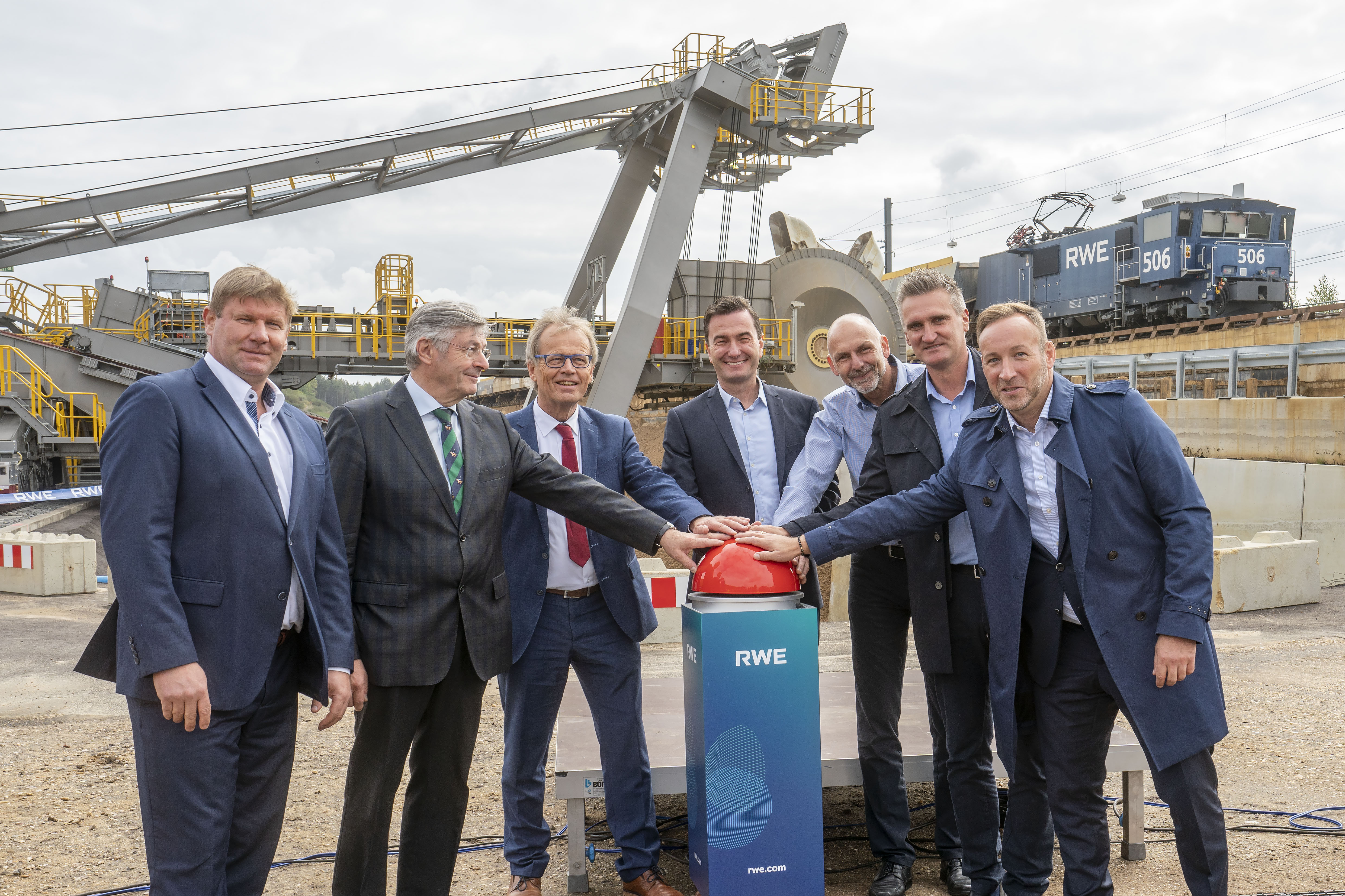 Eine Gruppe von Männern in Anzügen drückt einen großen roten Knopf an einem Industriestandort mit Maschinen und einem Zug im Hintergrund.