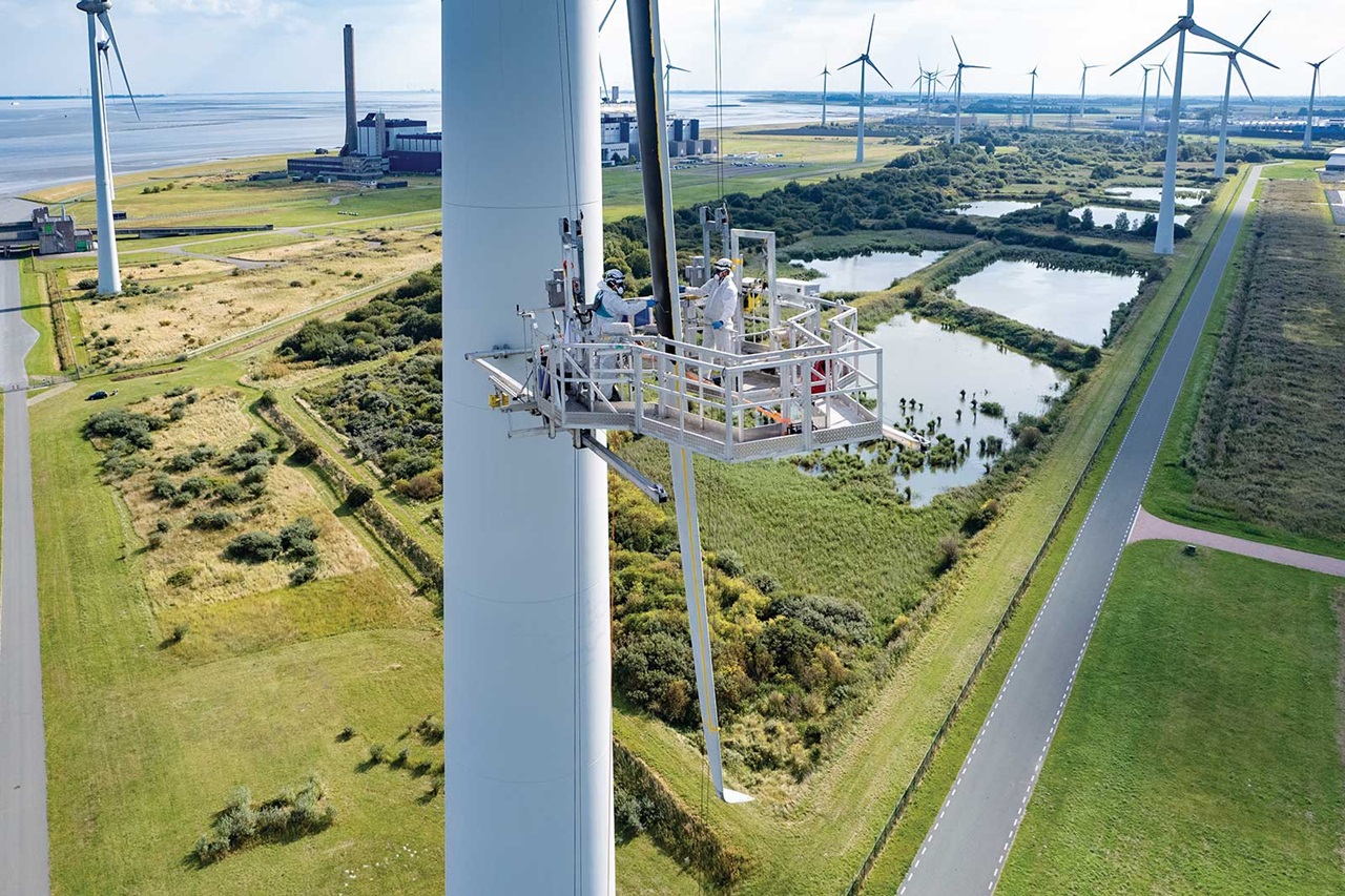 Eine Windkraftanlage wird von einer Plattform aus gewartet. Im Hintergrund sind weitere Turbinen und grüne Landschaften.
