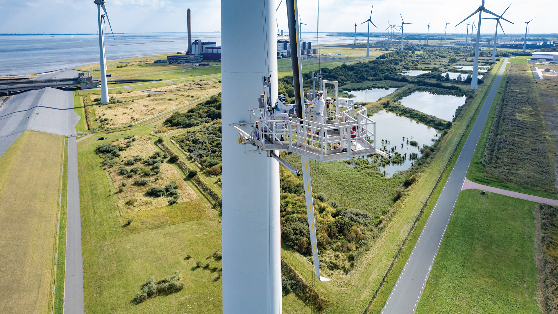 Eine Windkraftanlage wird von einer Plattform aus gewartet. Im Hintergrund sind weitere Turbinen und grüne Landschaften.