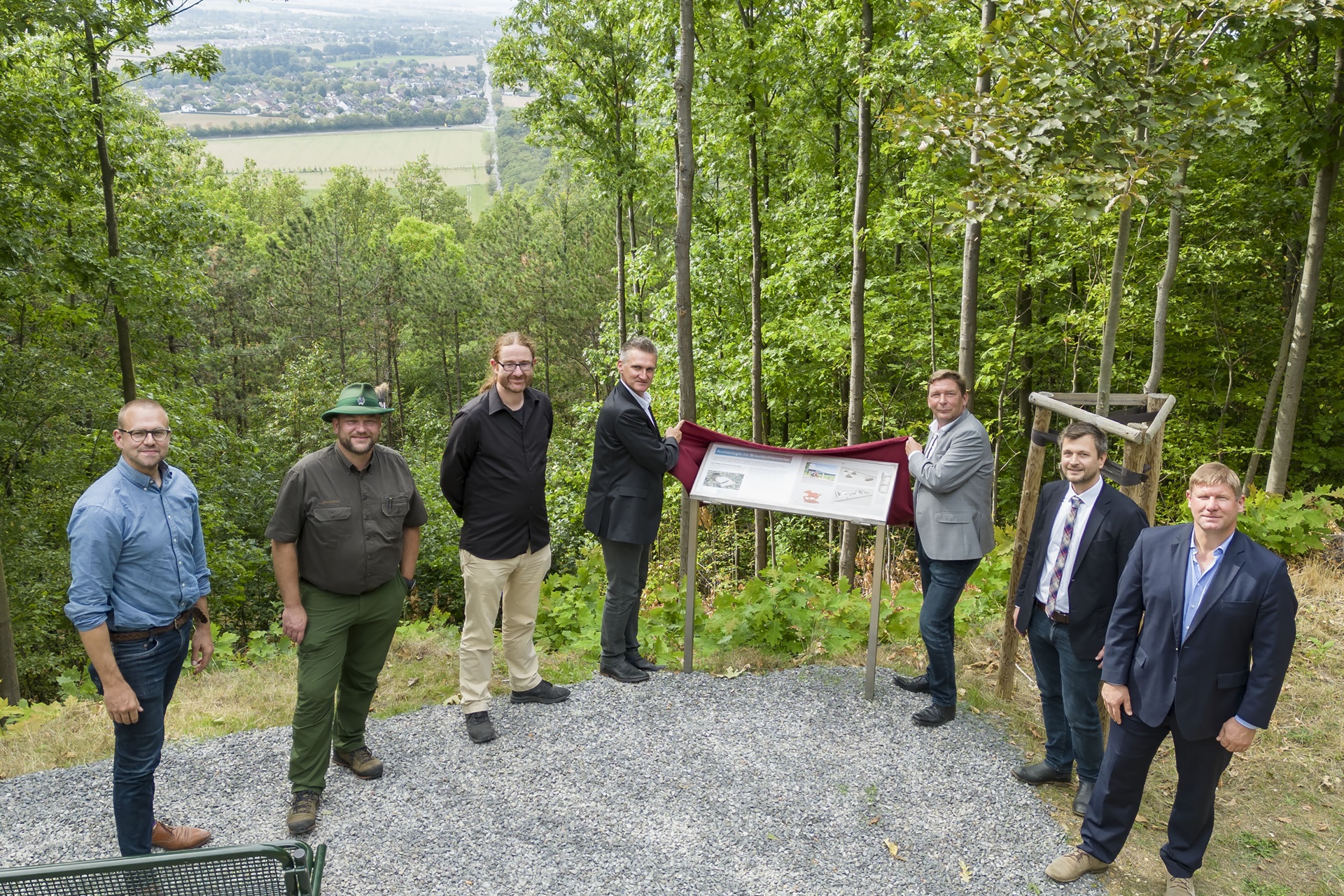 Eine Gruppe von Personen steht neben einem Schild in einem bewaldeten Gebiet mit einer malerischen Aussicht auf Felder und Hügel im Hintergrund.
