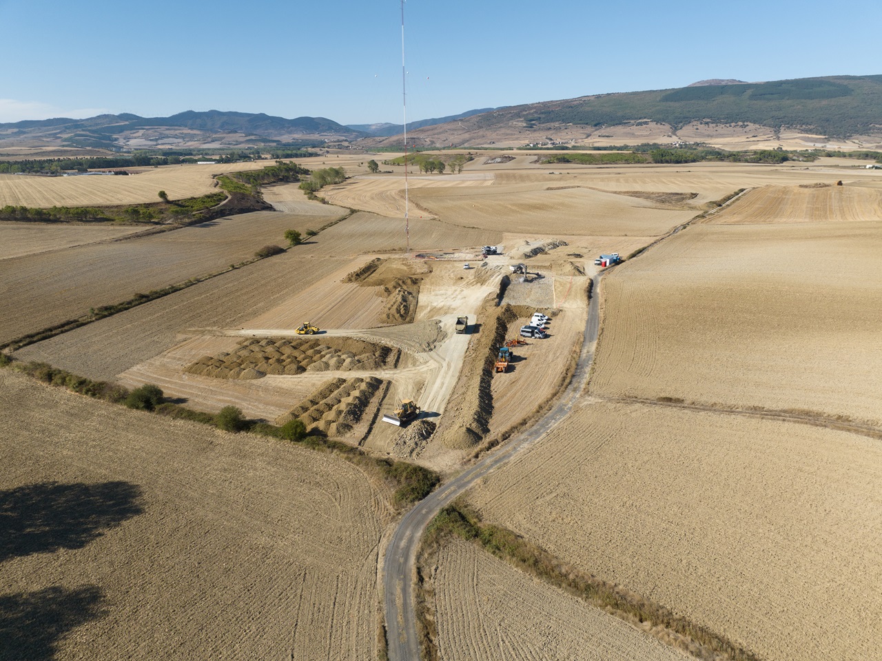 Eine Luftaufnahme einer Baustelle, umgeben von Feldern, mit schweren Maschinen und Fahrzeugen auf einem Schotterweg.