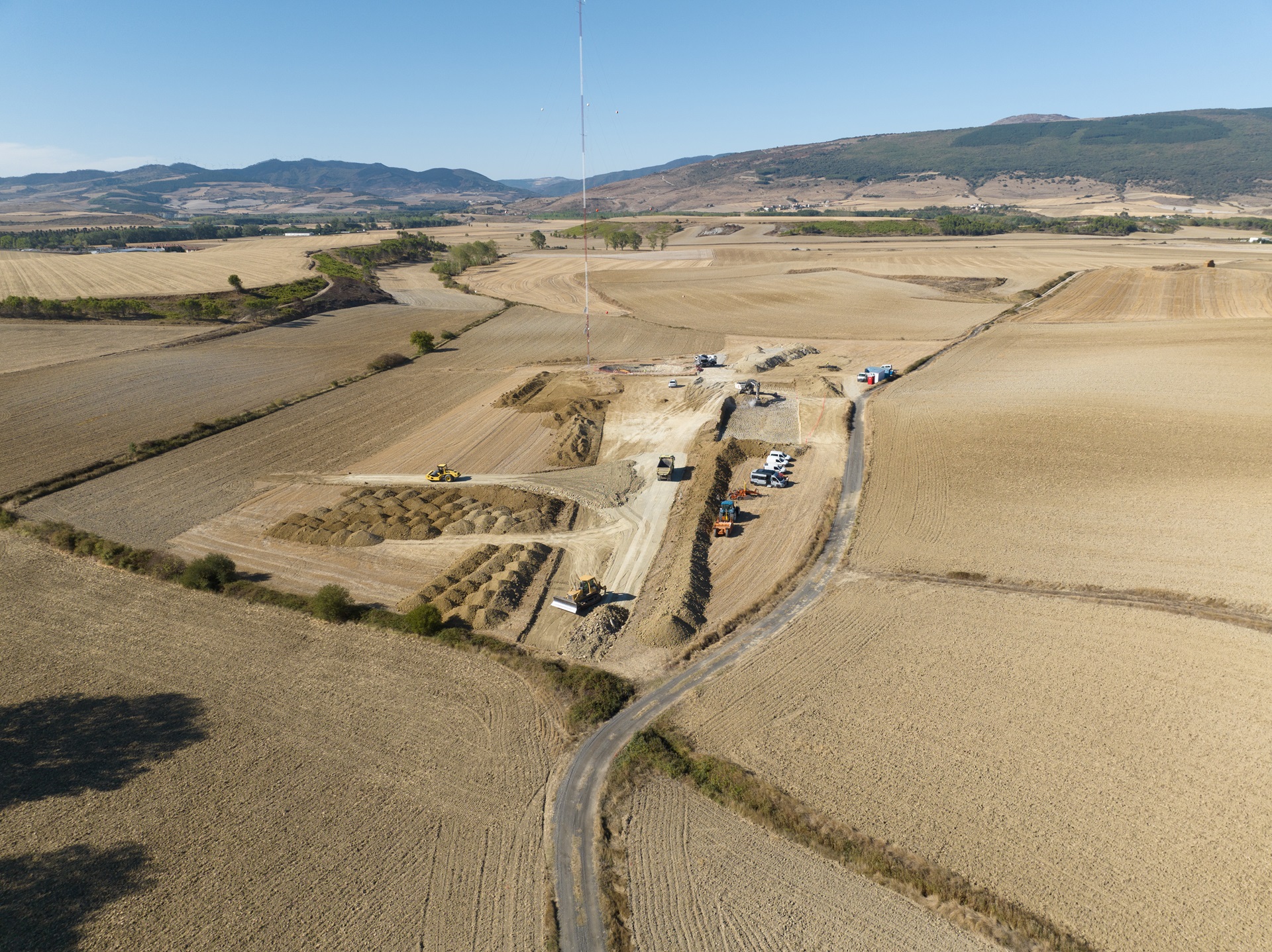 Eine Luftaufnahme einer Baustelle, umgeben von Feldern, mit schweren Maschinen und Fahrzeugen auf einem Schotterweg.