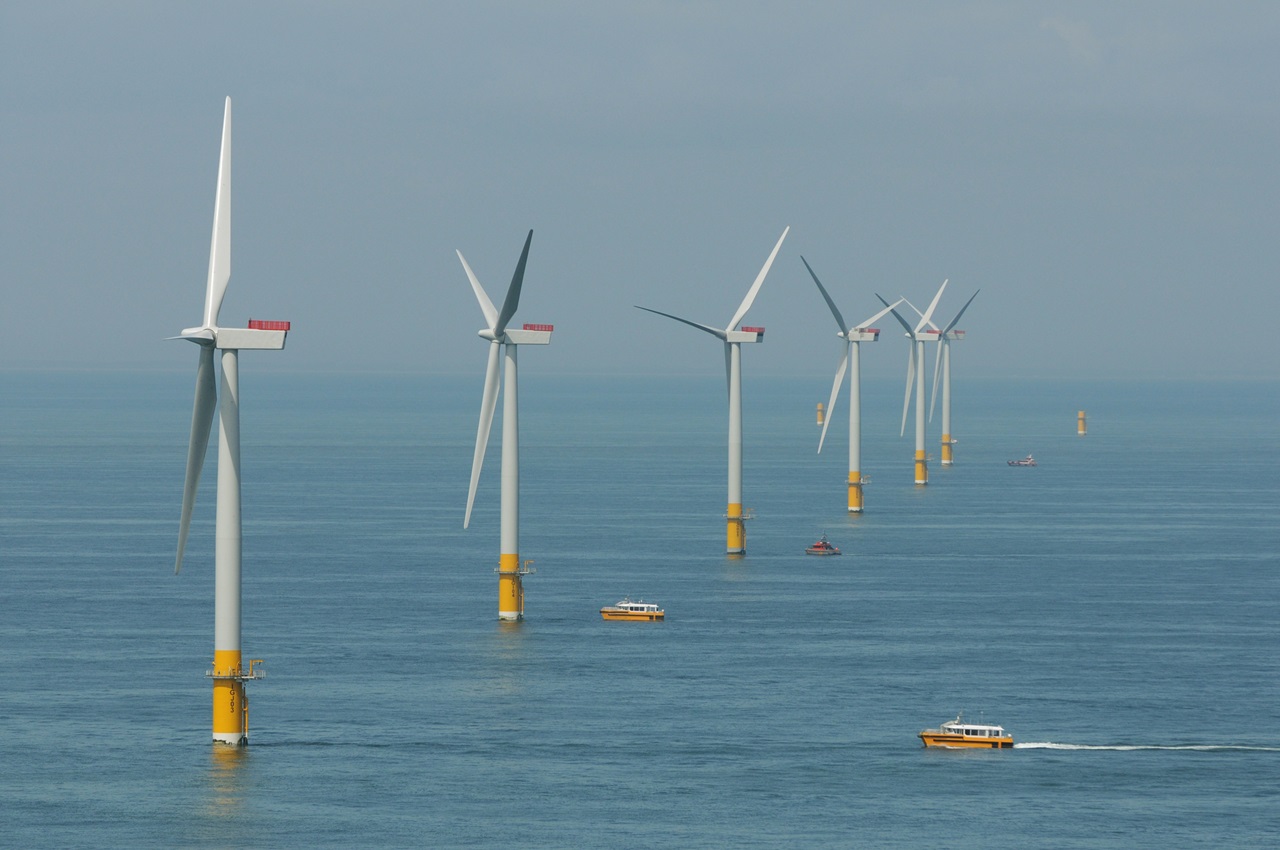 A row of wind turbines in the sea, with boats navigating nearby under a clear sky.