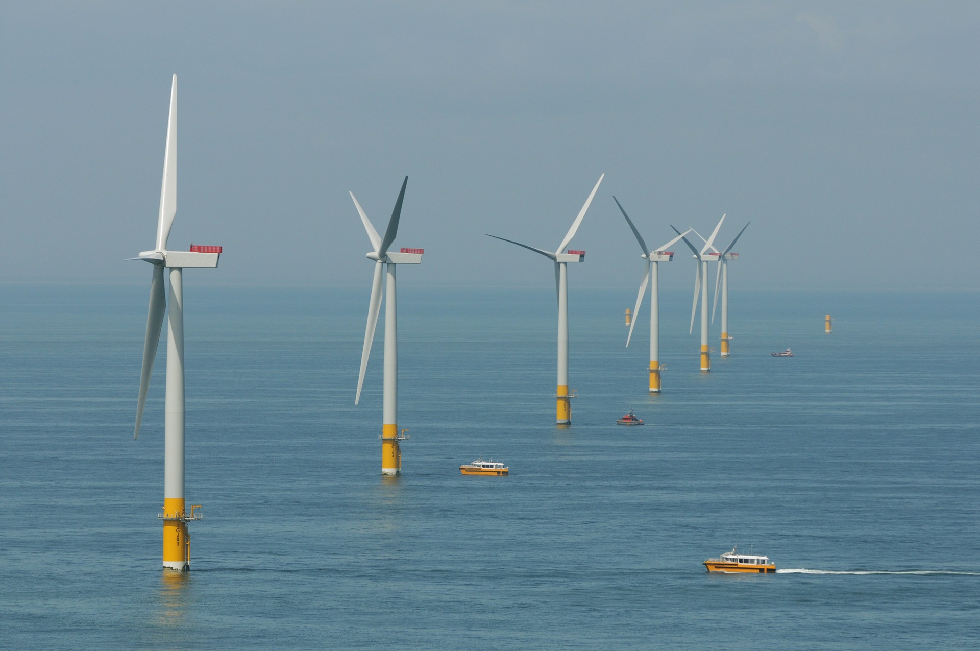 A row of wind turbines in the sea, with boats navigating nearby under a clear sky.