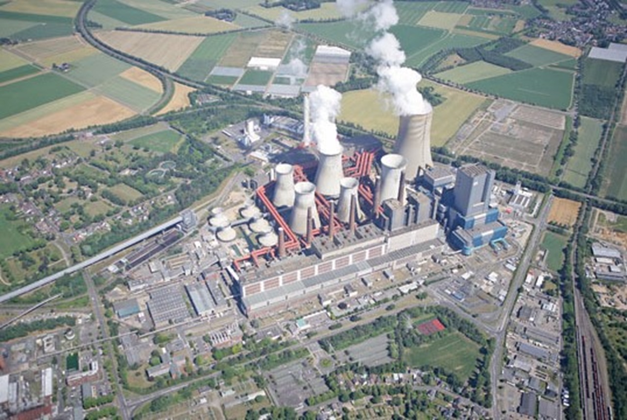 Aerial view of a power plant with cooling towers emitting steam, surrounded by green fields and roads.