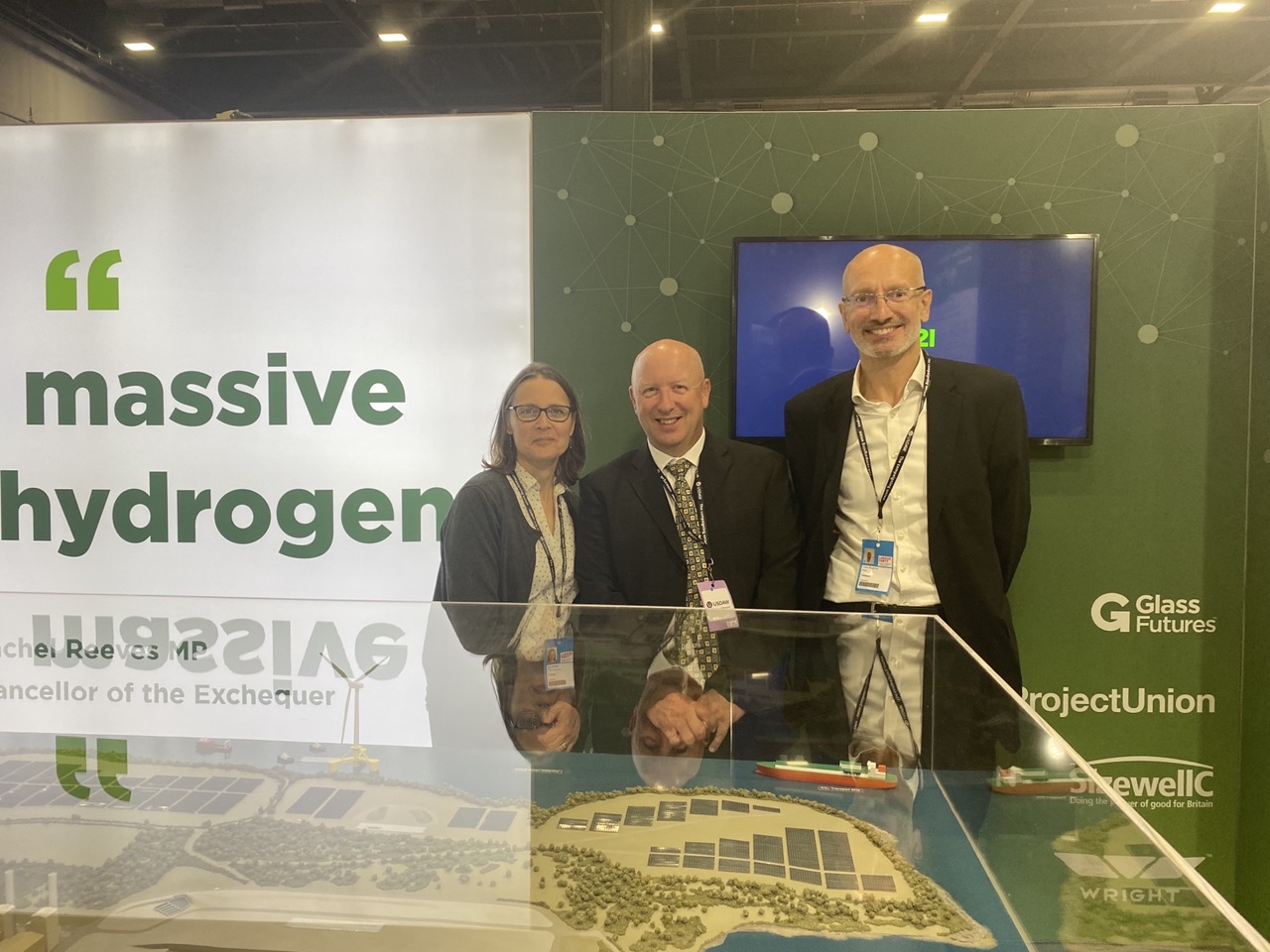 Three individuals pose in front of a large display featuring a 'massive hydrogen' sign at an event.