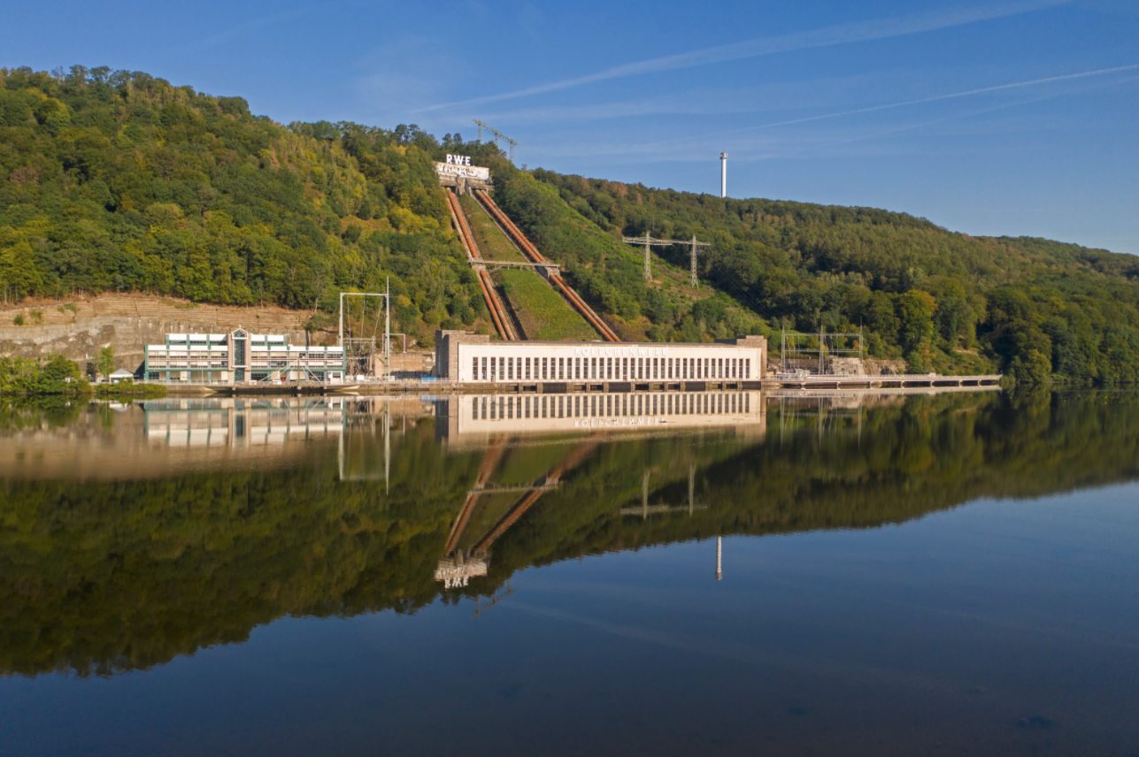 Ein Wasserkraftwerk mit Spiegelung im Wasser, umgeben von Bäumen und Hügeln unter einem klaren blauen Himmel.