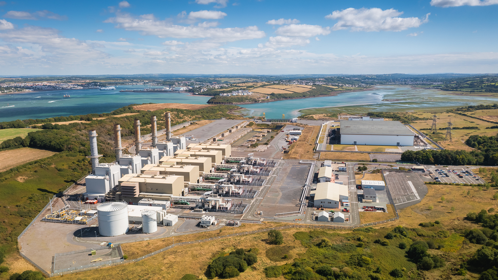 An aerial view of an industrial facility with gas turbines, storage tanks, and a river landscape.