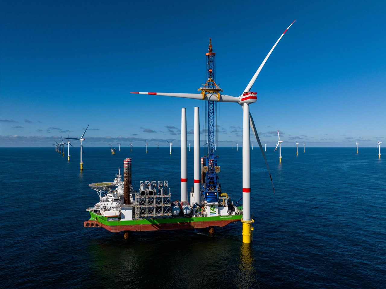 Ein großes Offshore-Windkraftinstallationsschiff mit mehreren Windturbinen im Hintergrund unter einem klaren blauen Himmel.