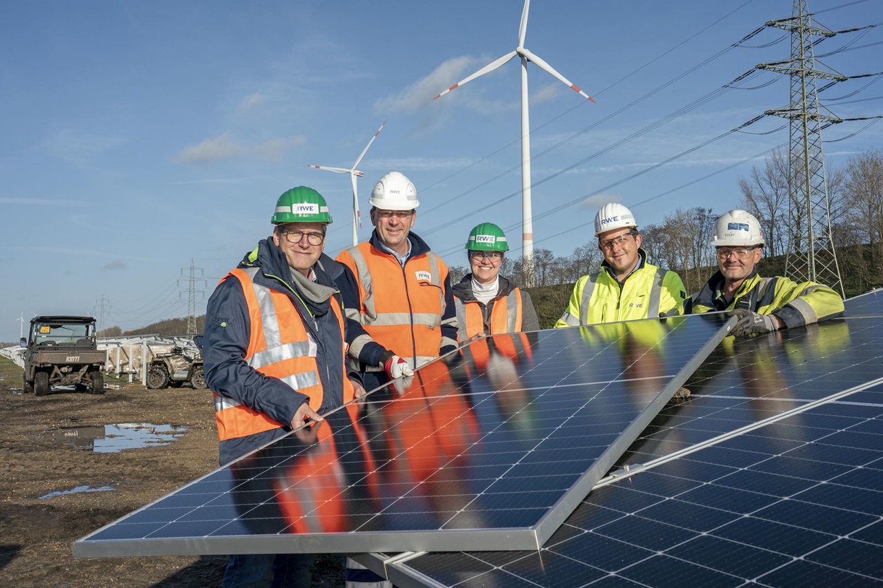 Eine Gruppe von fünf Arbeitern in Schutzkleidung inspiziert Solarpanels mit Windkraftanlagen im Hintergrund.