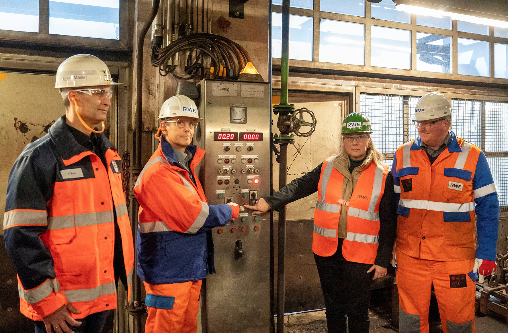 Vier Arbeiter in Schutzausrüstung interagieren mit einem Bedienfeld in einer Einrichtung und zeigen Teamarbeit in einem industriellen Umfeld.