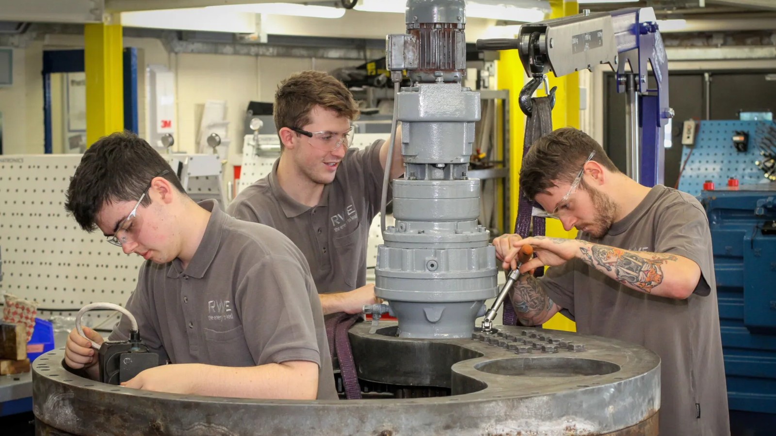 Three individuals work on mechanical equipment in a workshop, using tools and equipment on a large machine.