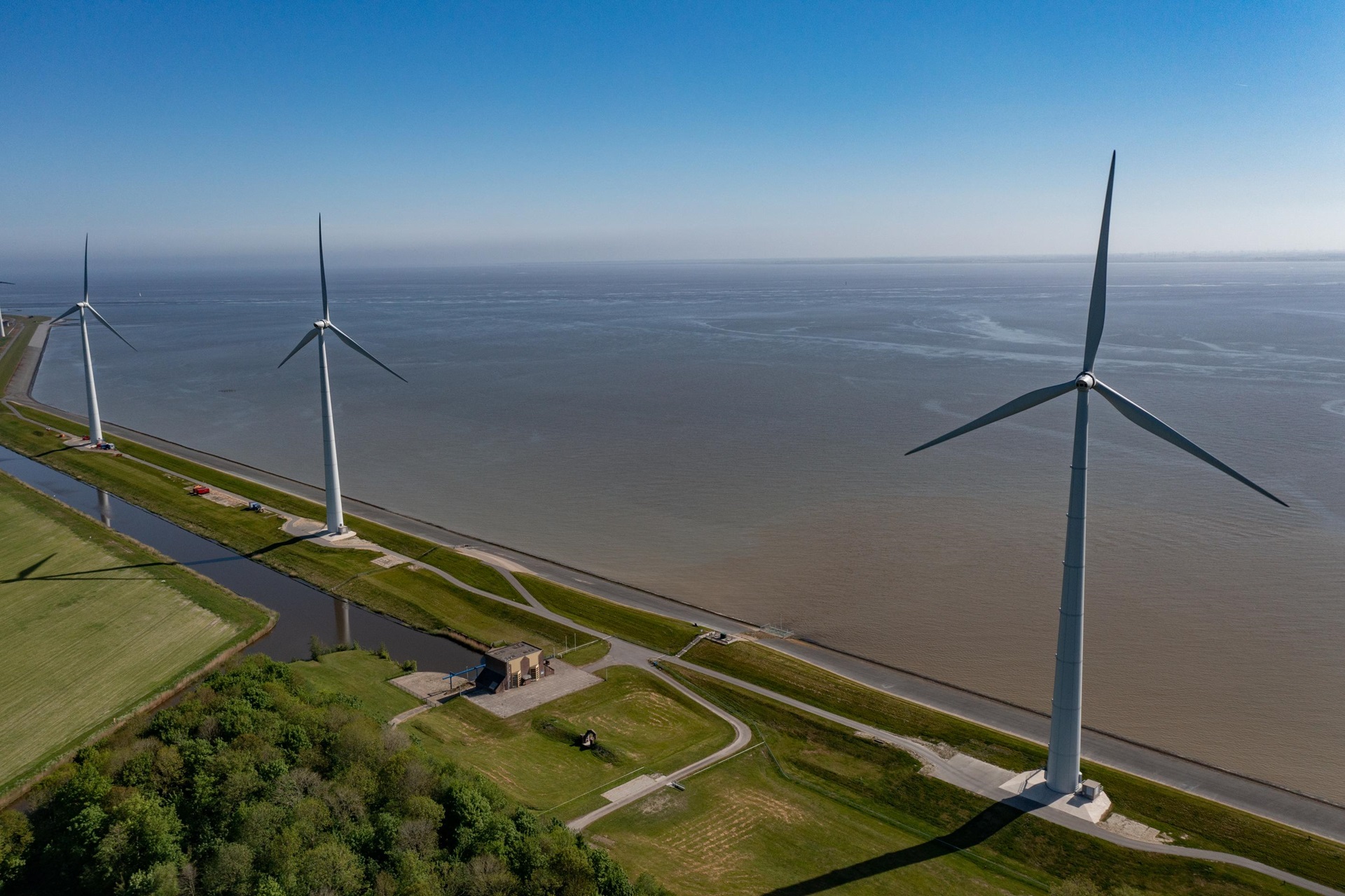 Aerial view of three wind turbines along a waterway with grassy banks and a small building nearby.