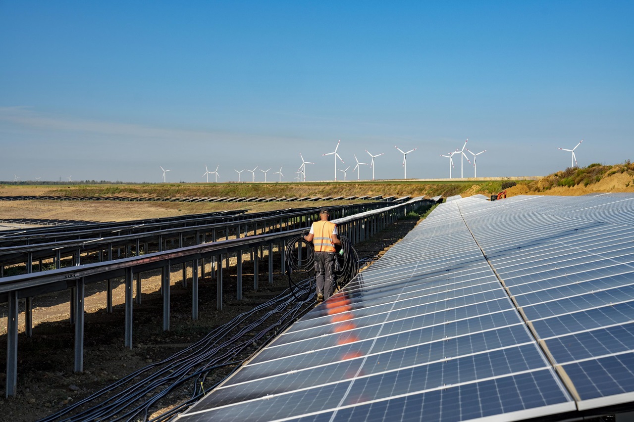 A worker in a high-visibility vest walks between solar panels with wind turbines in the background under a clear blue sky.