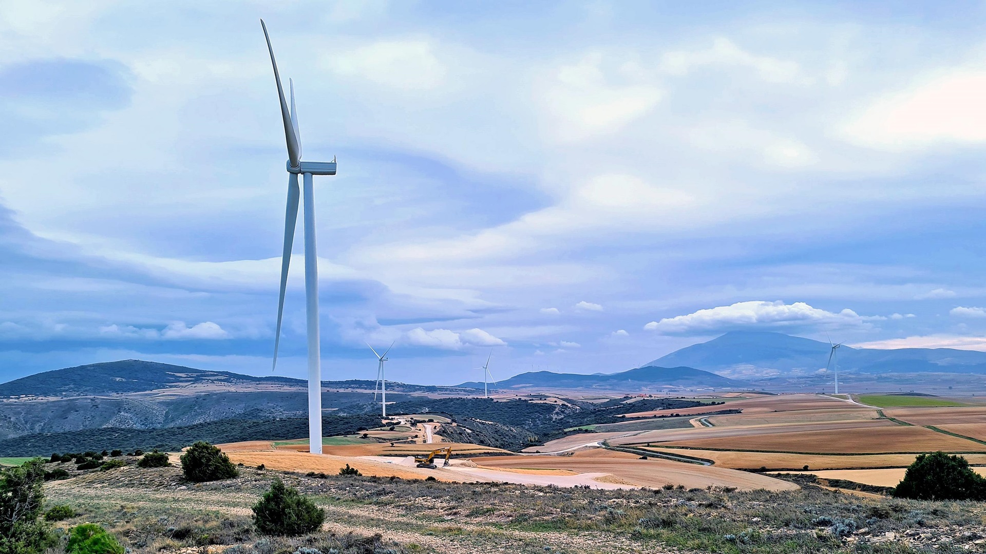 Eine Landschaft mit Windkraftanlagen vor einem bewölkten Himmel, mit sanften Feldern und fernen Bergen.