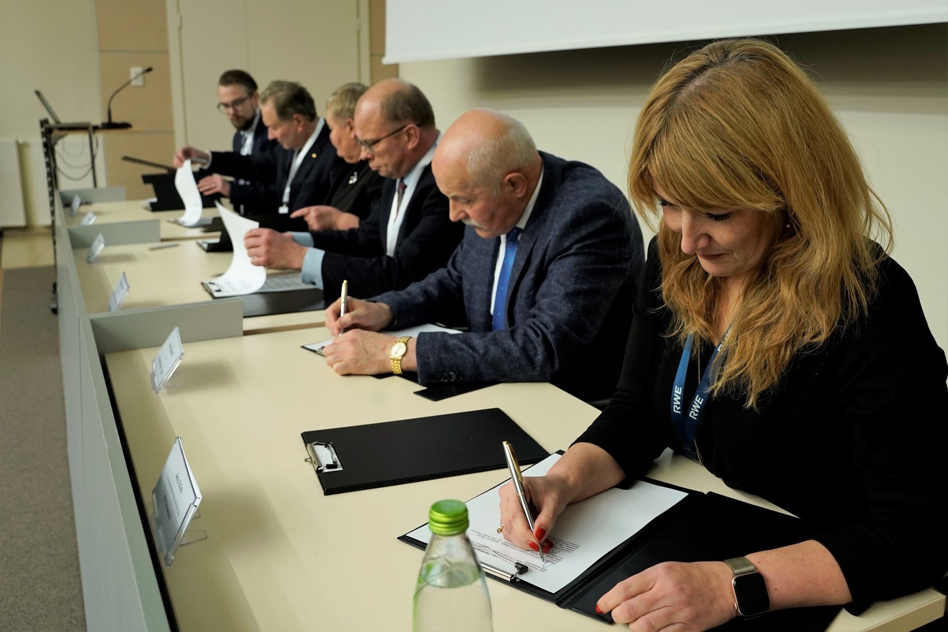 A panel of individuals seated at a table, engaged in writing and signing documents during a meeting.