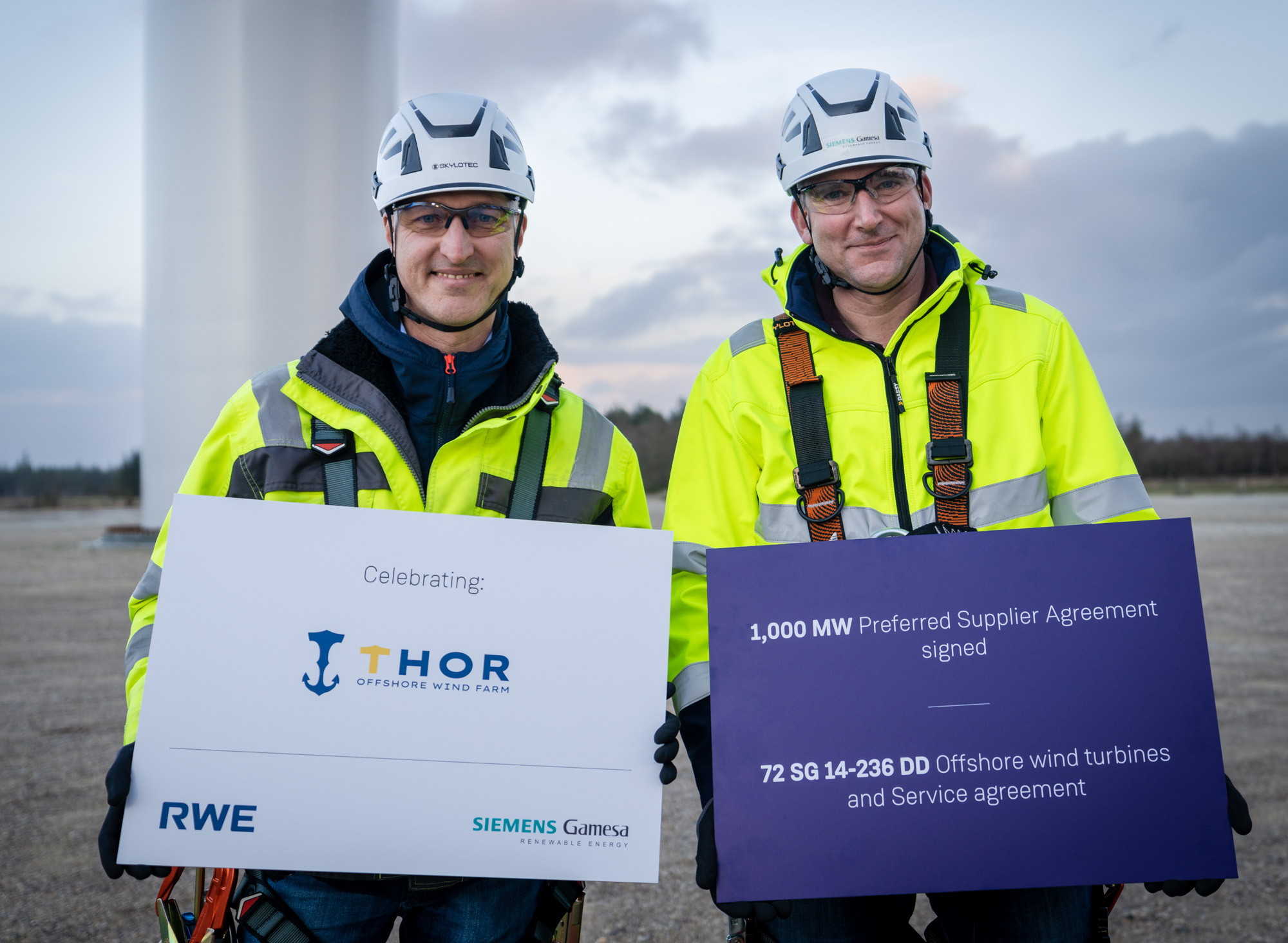Two workers celebrating the THOR Offshore Wind Farm agreement, holding signs about a preferred supplier agreement and wind turbines.