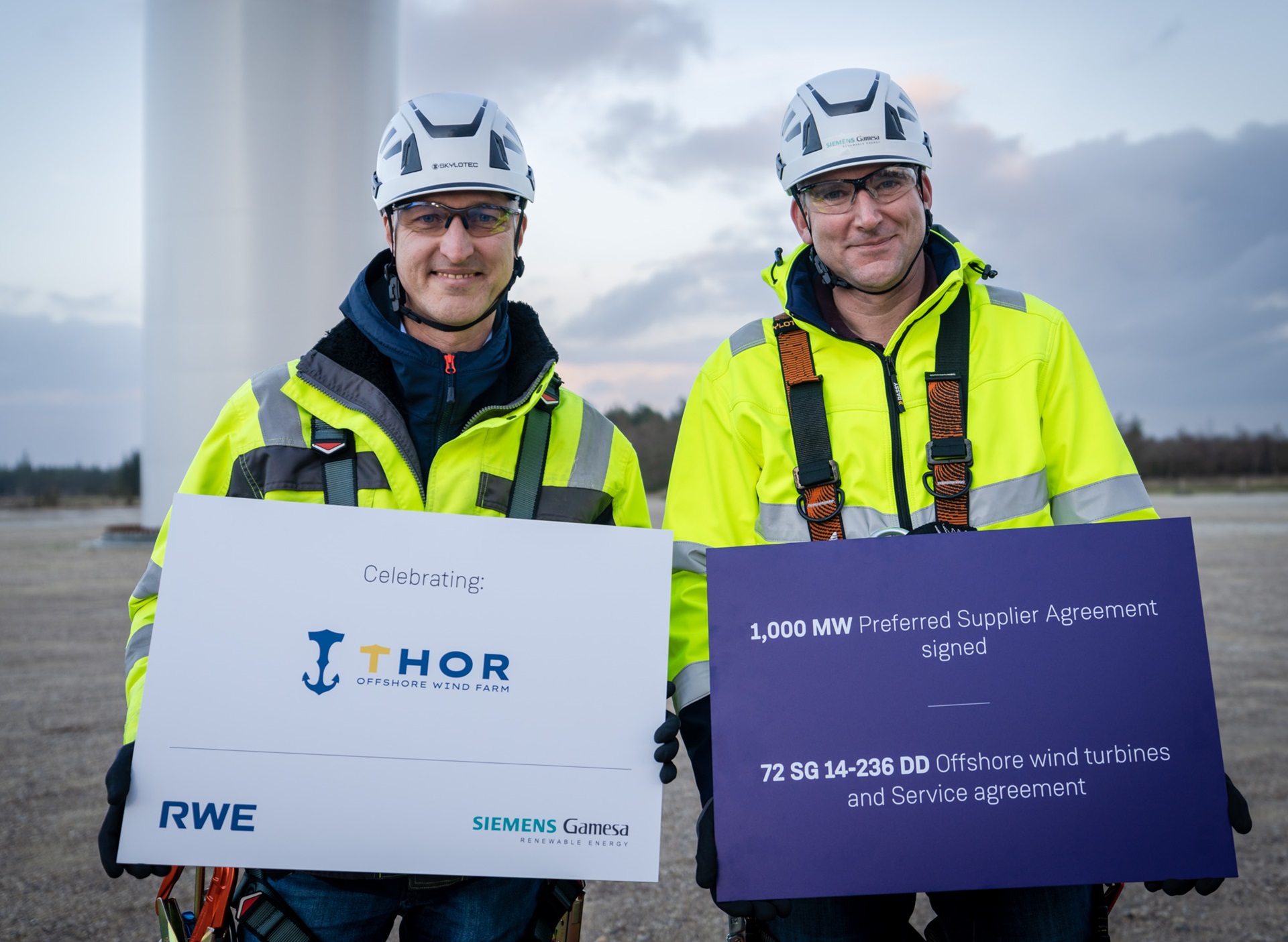 Two workers celebrating the THOR Offshore Wind Farm agreement, holding signs about a preferred supplier agreement and wind turbines.