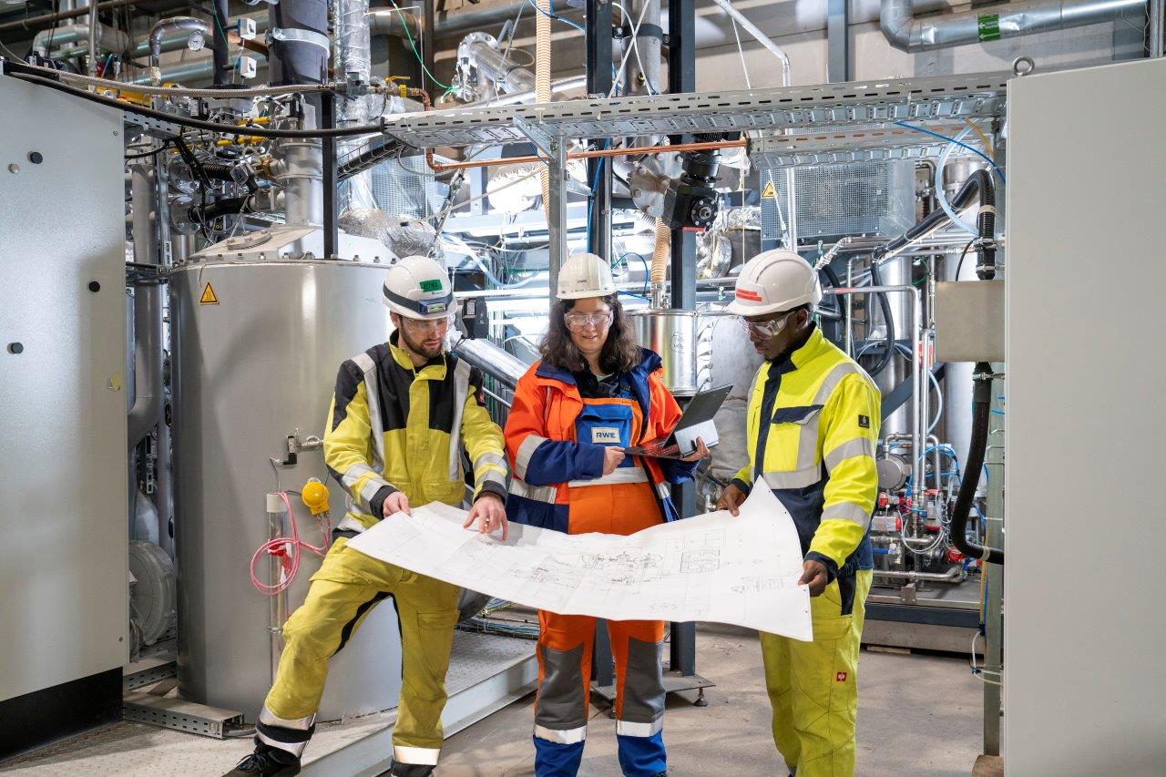 Drei Arbeiter in Sicherheitskleidung prüfen Pläne in einer industriellen Umgebung voller Maschinen und Rohre.