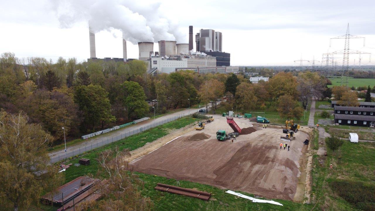 Eine Luftaufnahme von Baumaschinen, die auf einer Baustelle arbeiten, mit einem Kraftwerk und Bäumen im Hintergrund.