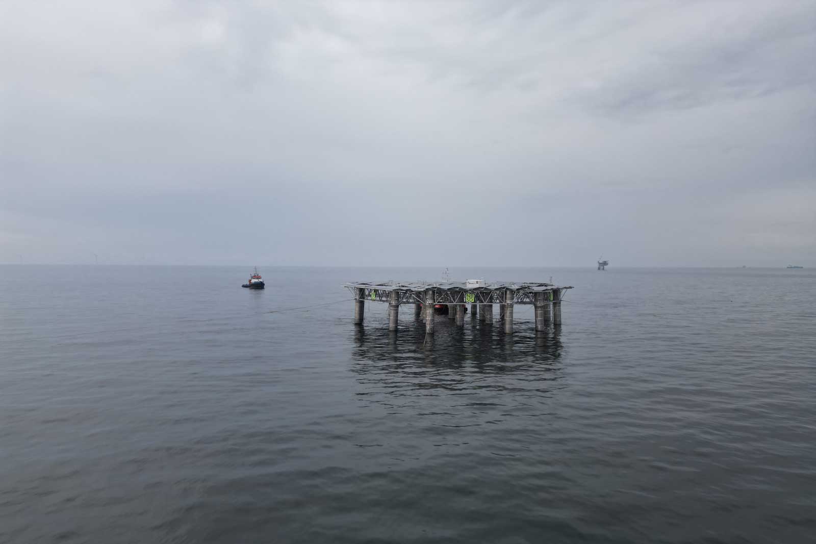 Eine Marineplattform steht in ruhigem Wasser, mit einem kleinen Boot in der Nähe und fernen Strukturen am Horizont unter einem bewölkten Himmel.