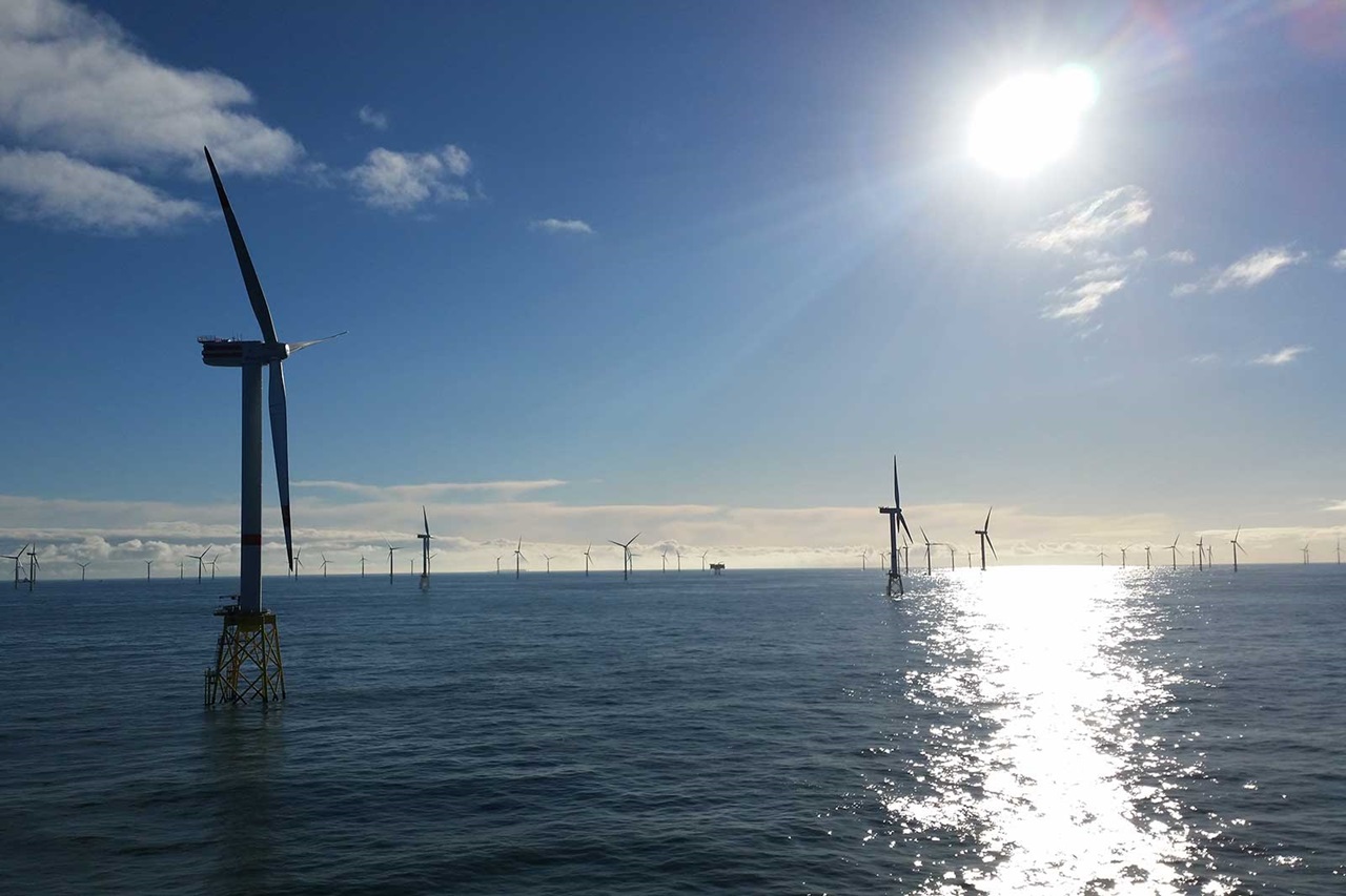 Several wind turbines stand in the water under a clear sky, while the sun shines brightly.