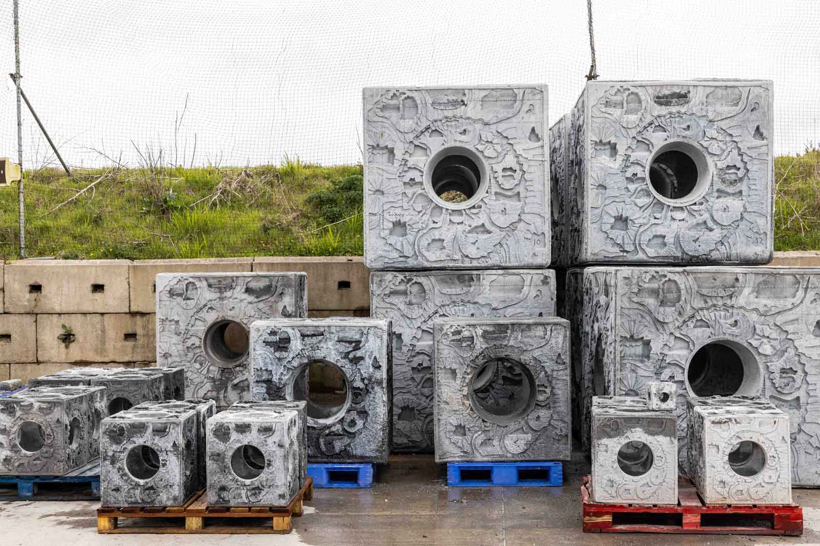 A collection of large concrete blocks with circular holes, stacked on blue and brown pallets, set against a grassy background.