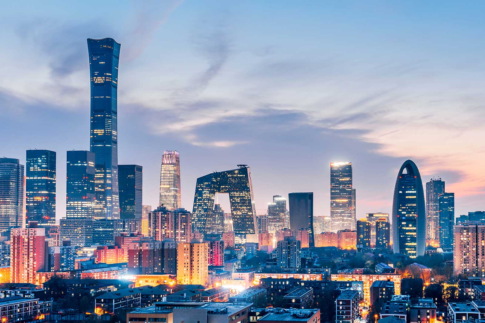 Illuminated midrise buildings occupy the foreground while glossy modern skyscrapers with varied silhouettes rise against a pastel blue and pink dusk sky.