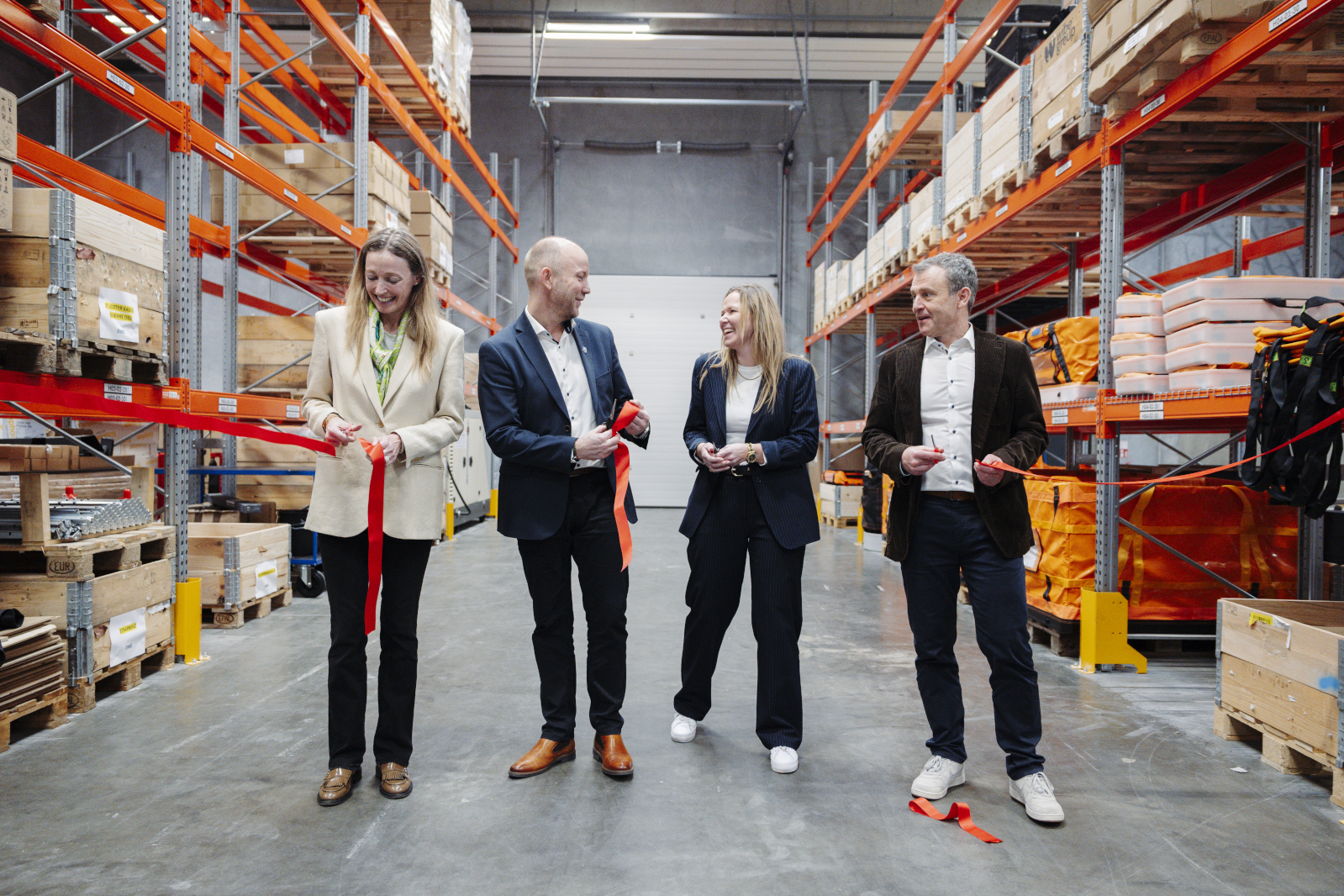 Four people stand in a warehouse for a ribbon-cutting ceremony, each holding a red ribbon to be cut.