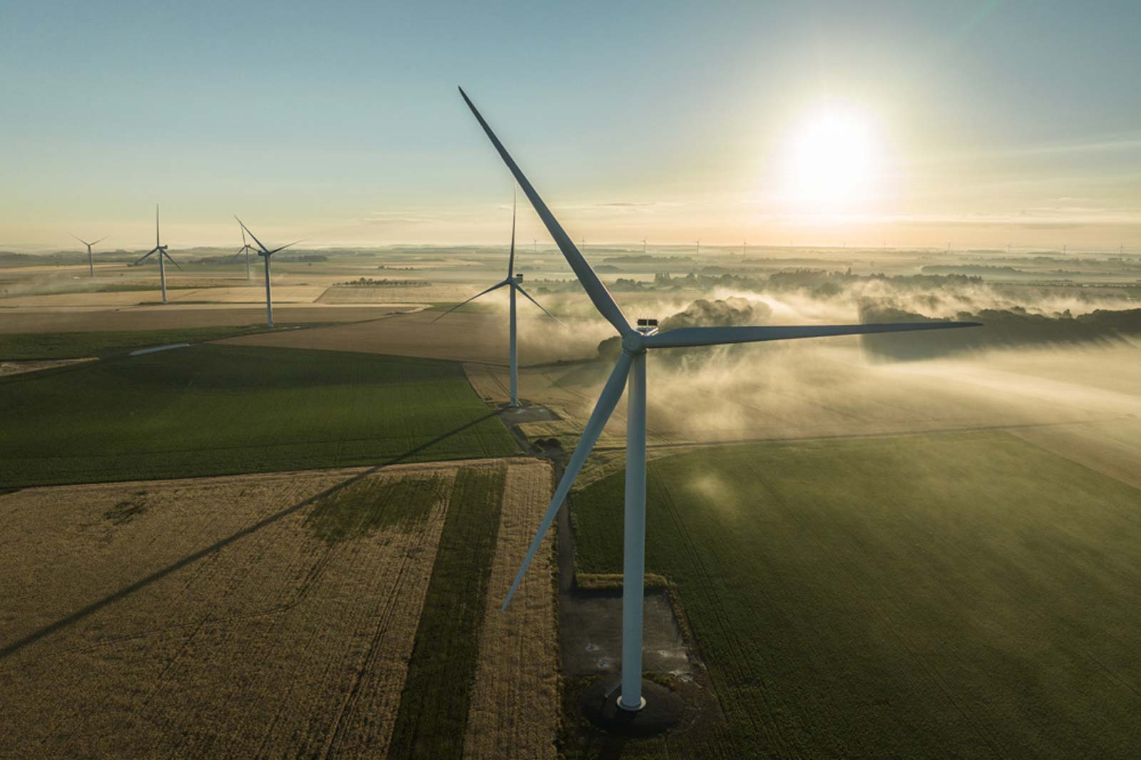 Aerial view of wind turbines in a lush green landscape at sunrise, with fog gently rising over the fields.
