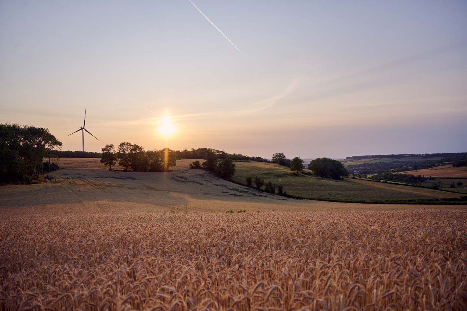 A serene landscape featuring golden wheat fields, a wind turbine, and a sunset over rolling hills.